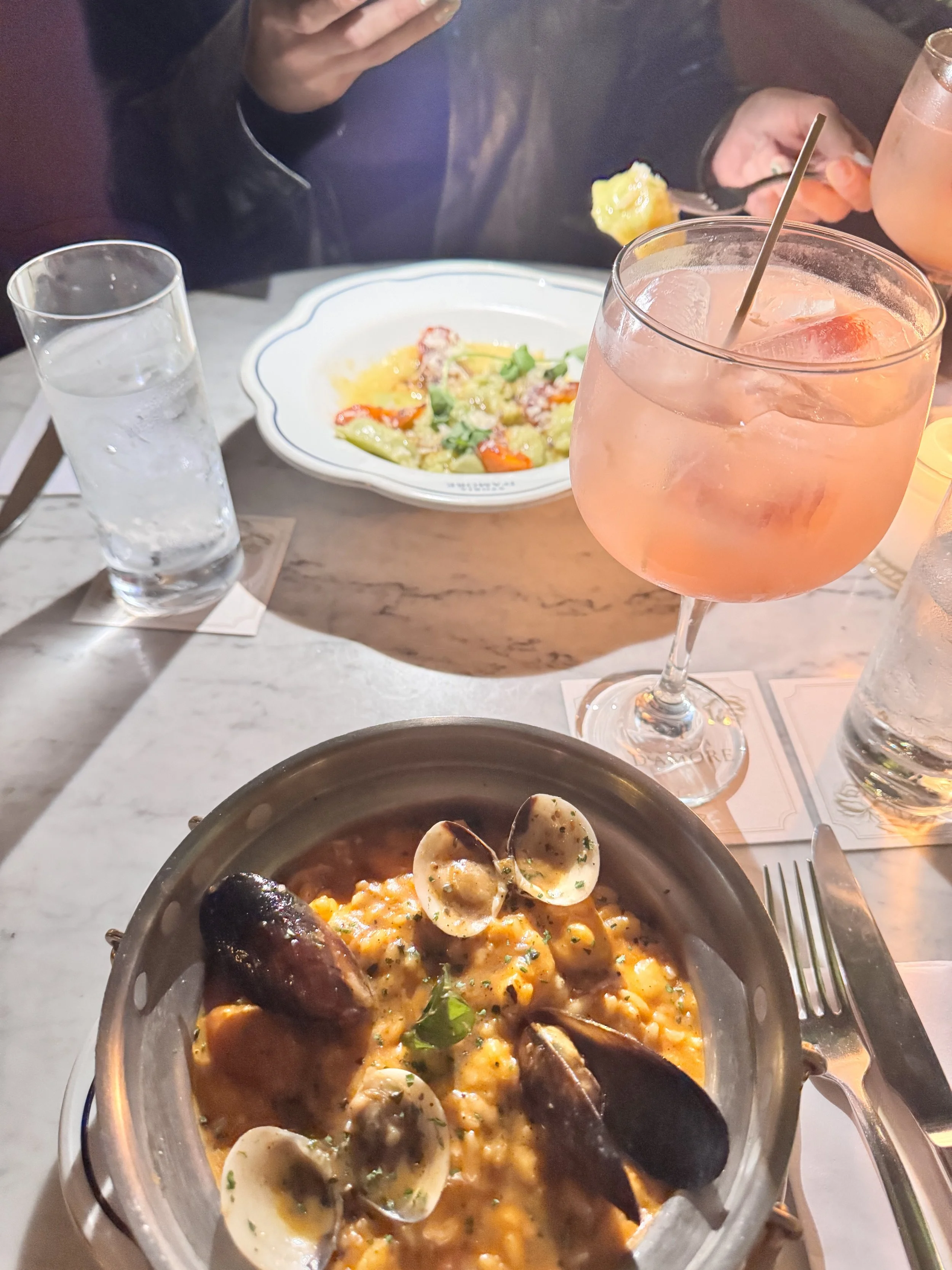 A table set with seafood risotto in a skillet, two glasses of pink cocktails, a glass of water, a salad on a white plate, and partially visible hands holding a fork with lemon in the background.