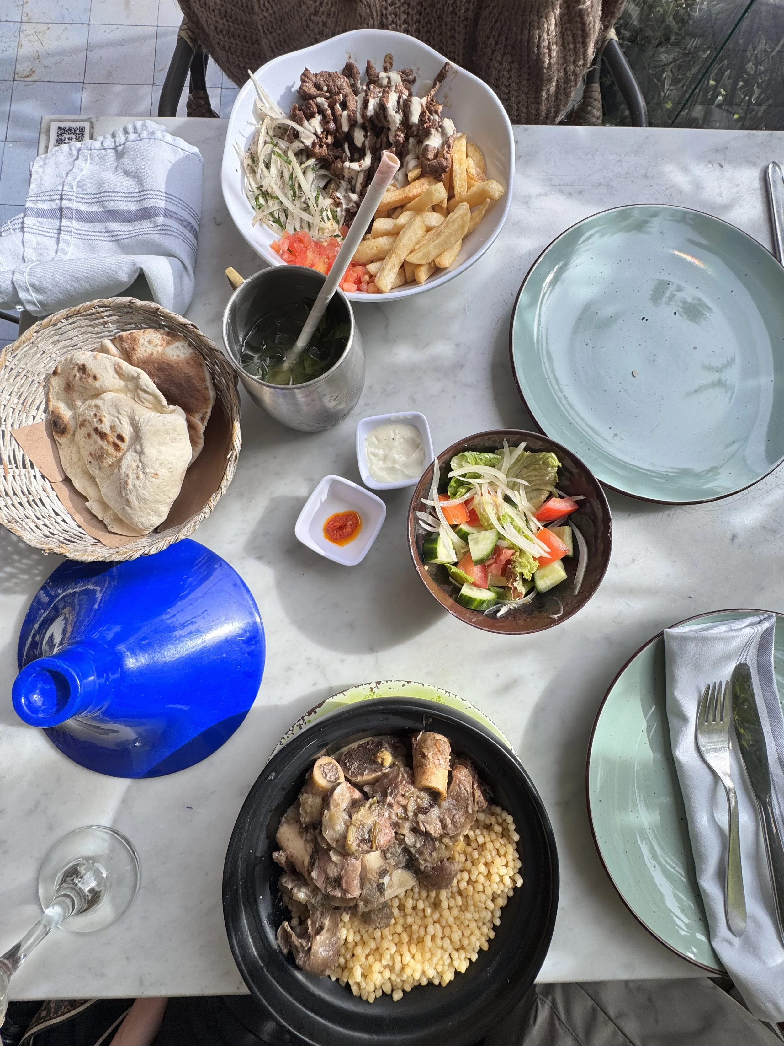 A table with various Middle Eastern dishes including a bowl of lamb and couscous, a basket of flatbread, a small salad, and a large bowl of beef and vegetable stew, along with drinks, condiments, and dining utensils.