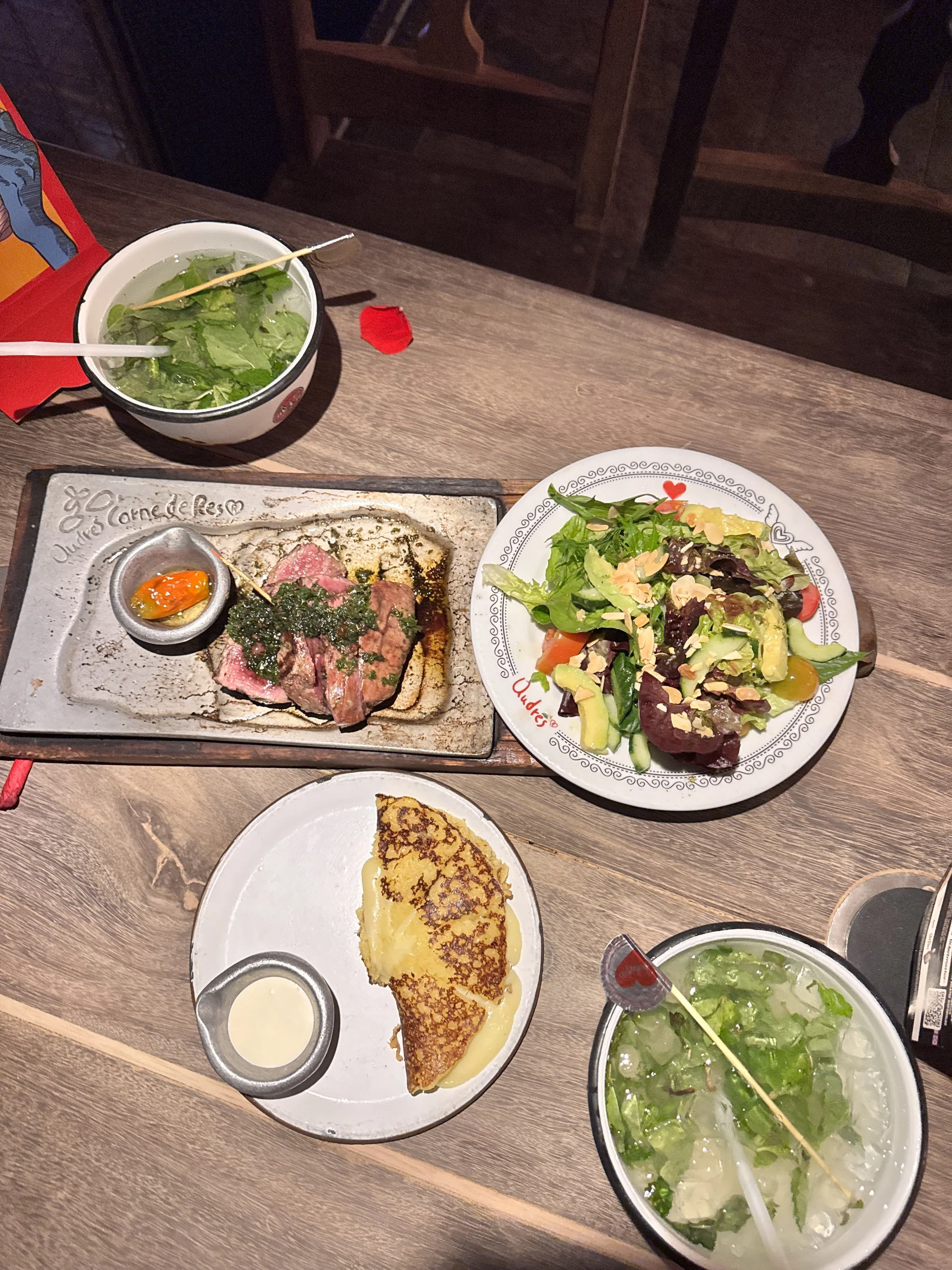 A dinner table with two bowls of green salad, a plate of sliced meat with sauce, a crepe with a side of cream, and two glasses of water, set on a wooden table.