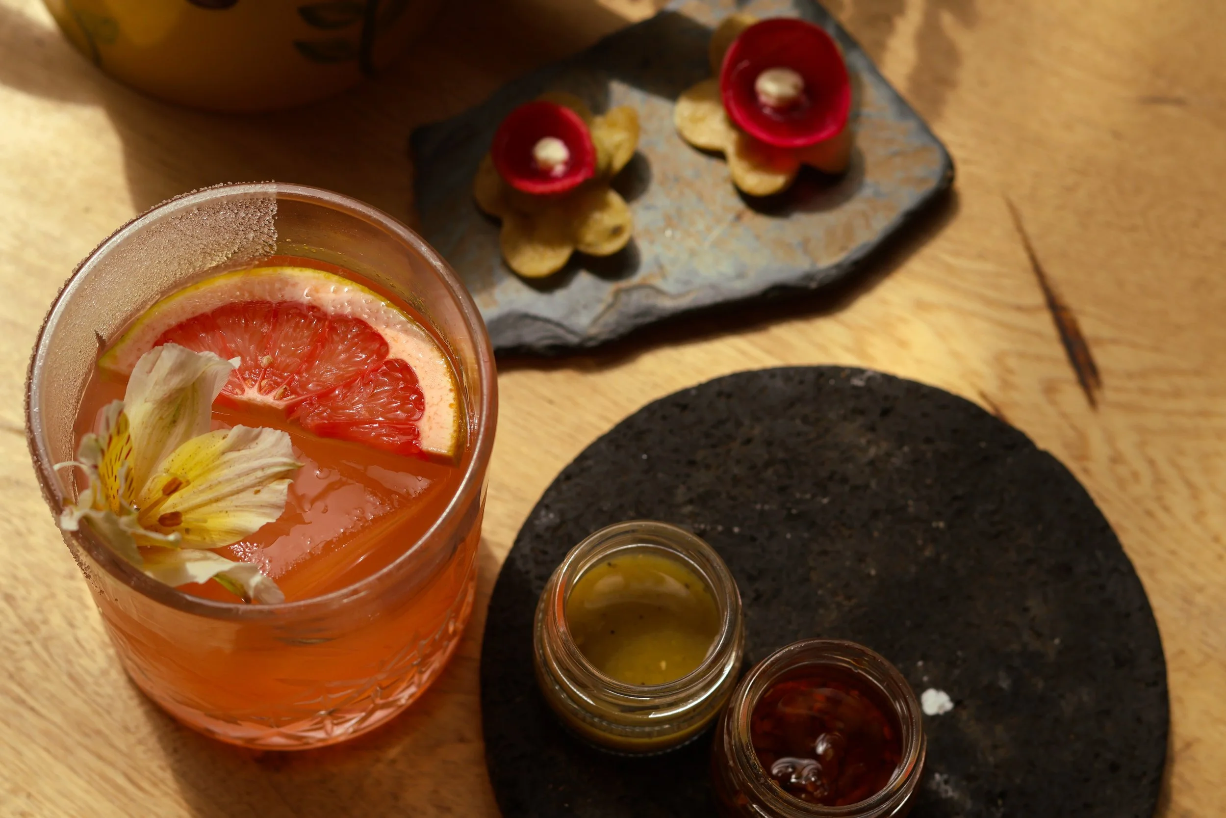 A drink with a grapefruit slice and flower, two small jars of sauces or condiments, and a plate with small appetizers topped with red flowers on a stone serving dish.