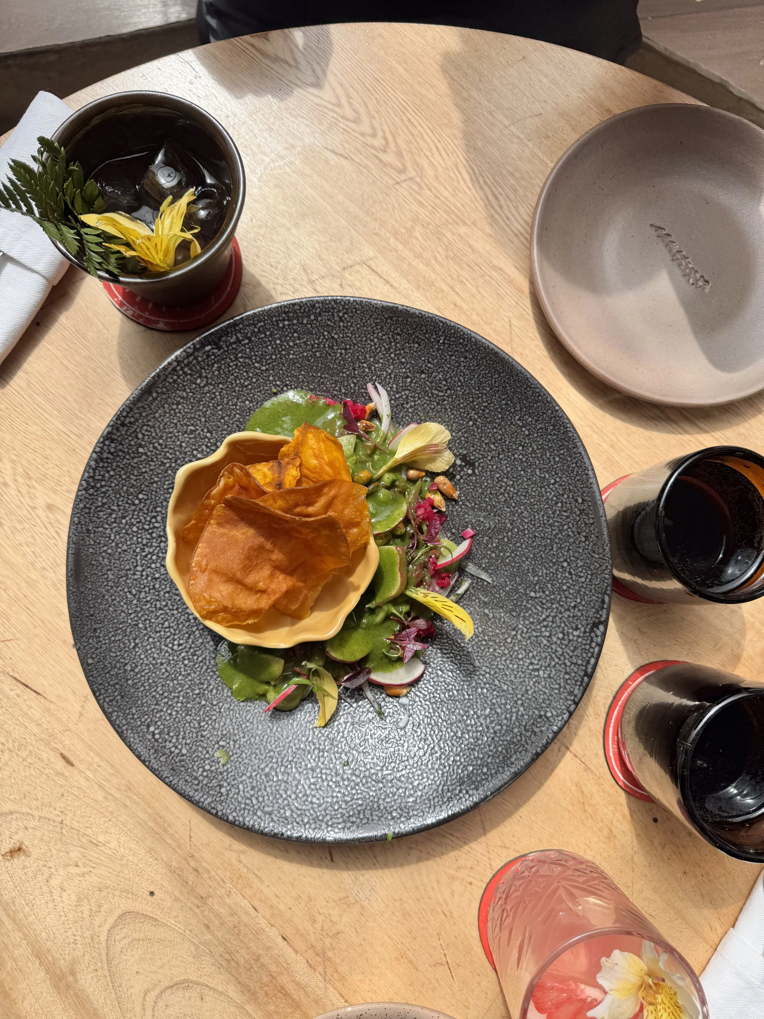A black plate with a salad containing sliced radishes, greens, and edible flowers, topped with a yellow bowl of crispy plantain chips. Surrounding the plate are glasses and cups with drinks on a wooden table.