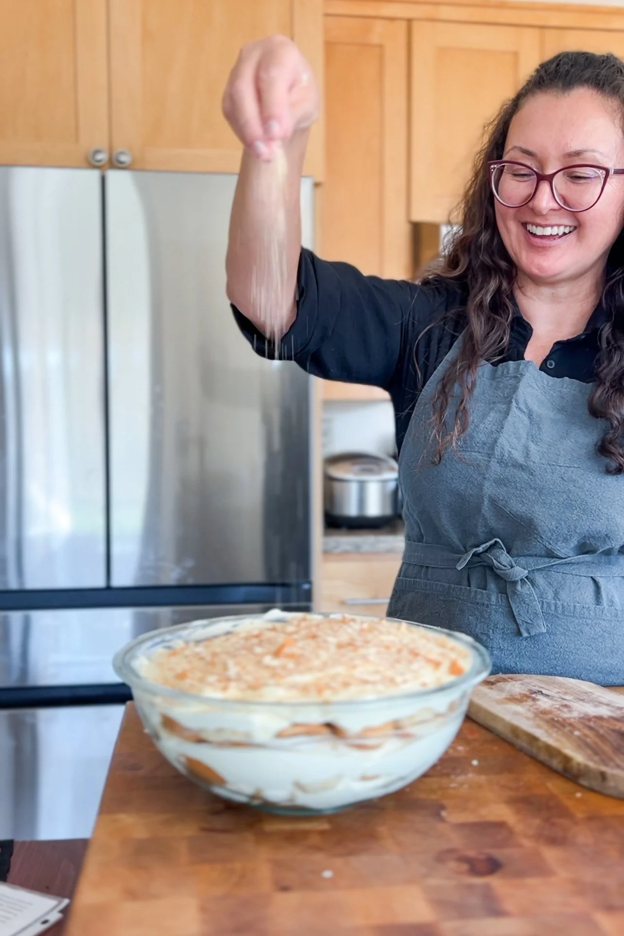 A woman with glasses and curly hair, smiling, wearing a black shirt and a gray apron, sprinkling cookie crumbs while baking in a kitchen. There is a pie dish on the countertop and a notebook nearby.