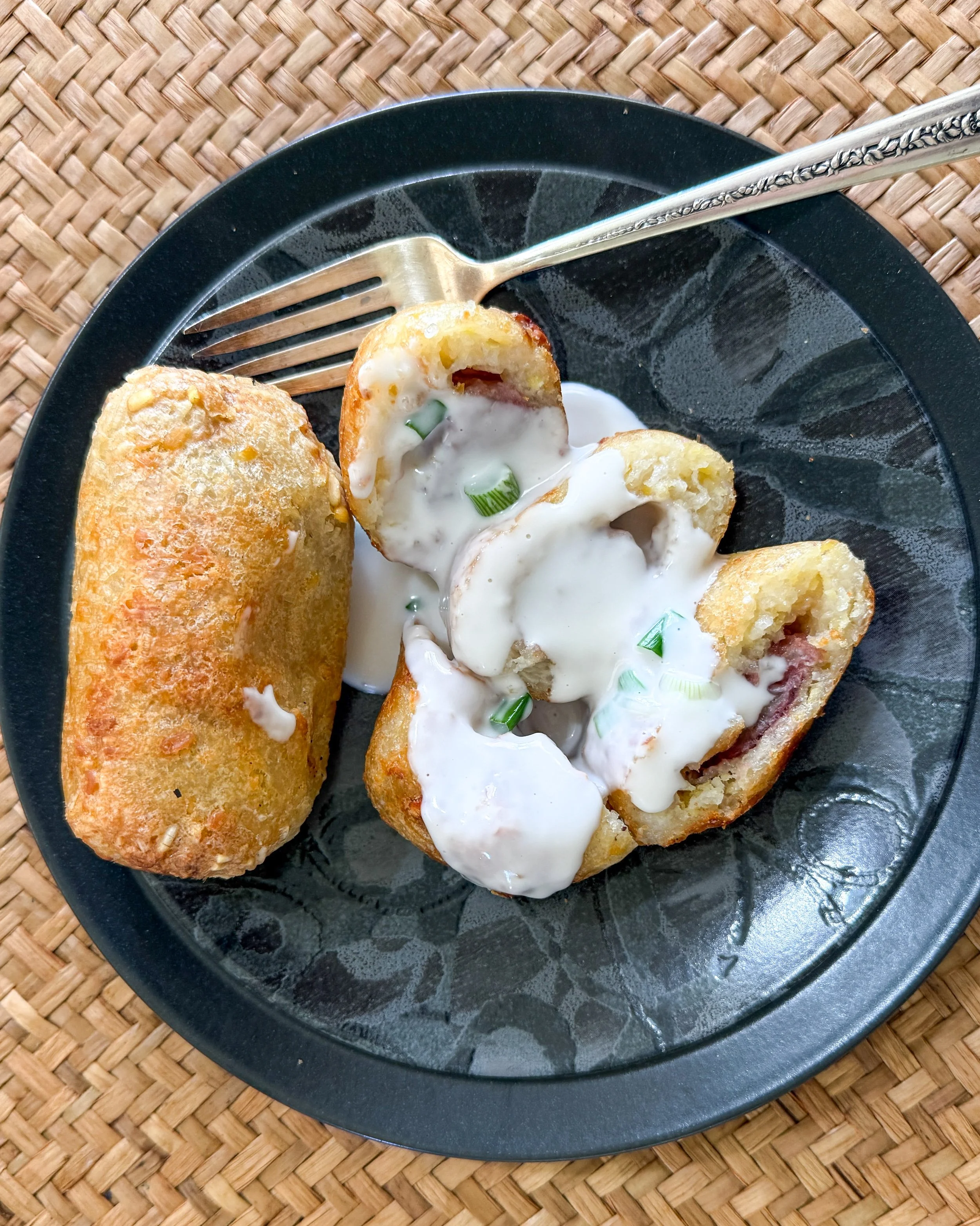 Crispy grilled sweet rice stuffed with bananas, topped with creamy coconut sauce and chopped green onions, served on a black plate.