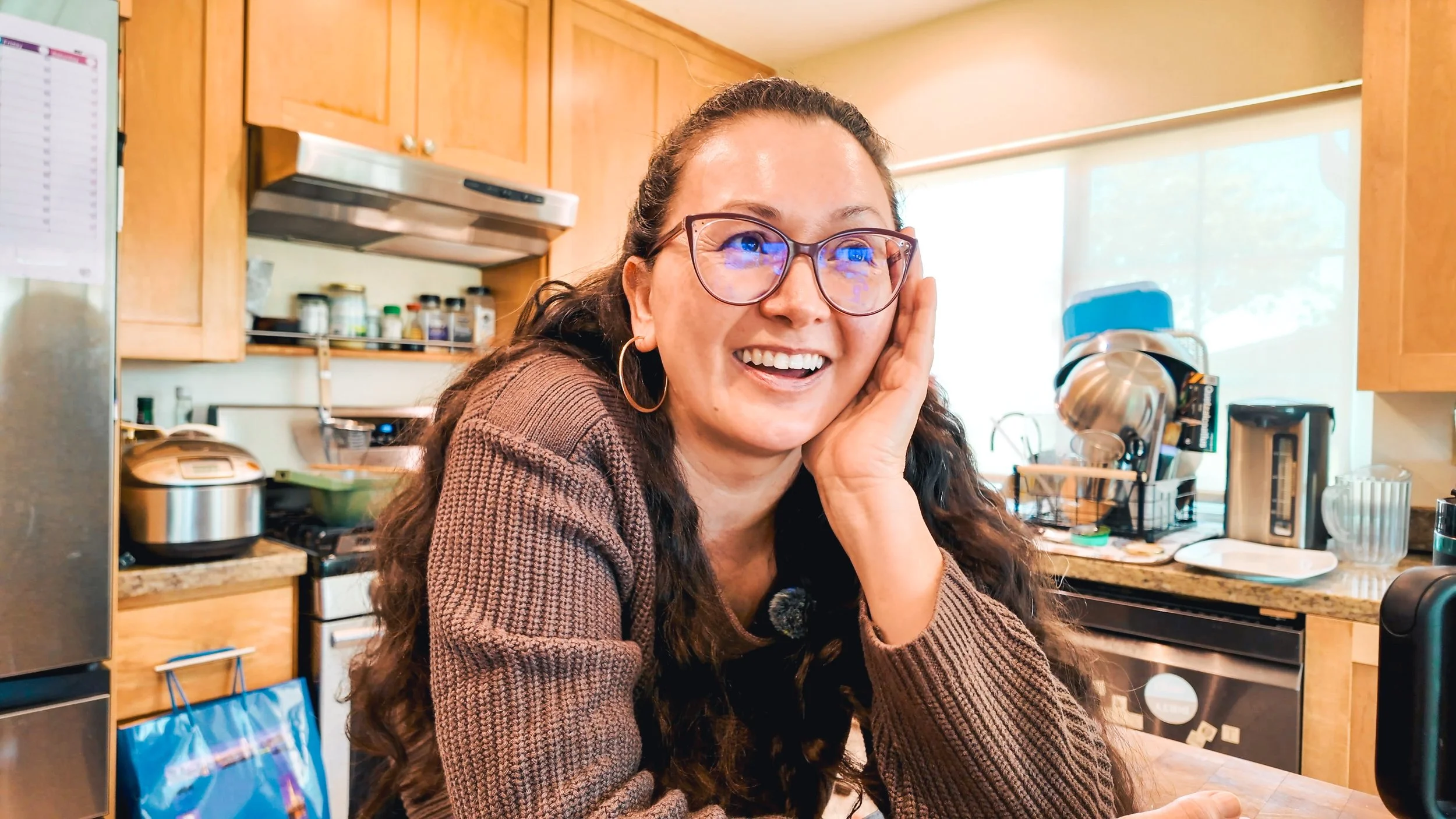 A woman with long curly hair and glasses smiling with her head resting on her hand in a kitchen.
