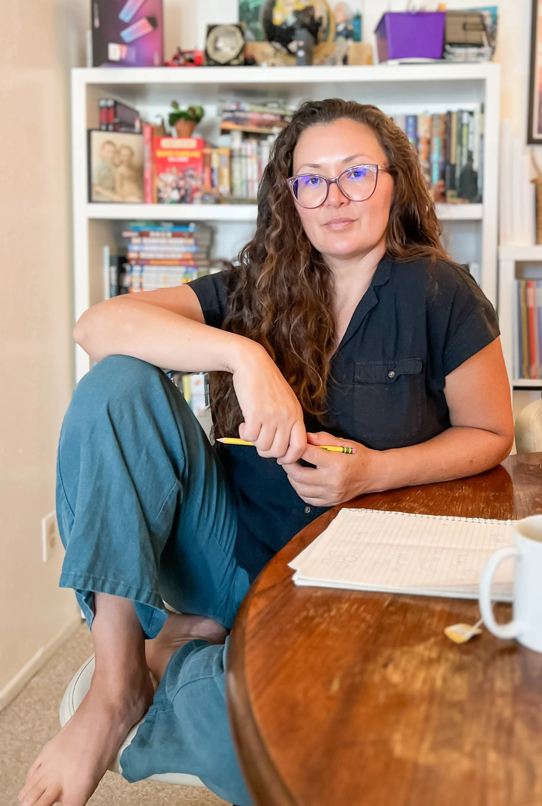 A woman with curly brown hair and glasses sitting at a wooden table with a young person leaning against her leg. The woman is smiling softly and holding a yellow pencil. There is a notebook with hand-drawn shapes on the table. Books and decorative items are on a white bookshelf in the background.