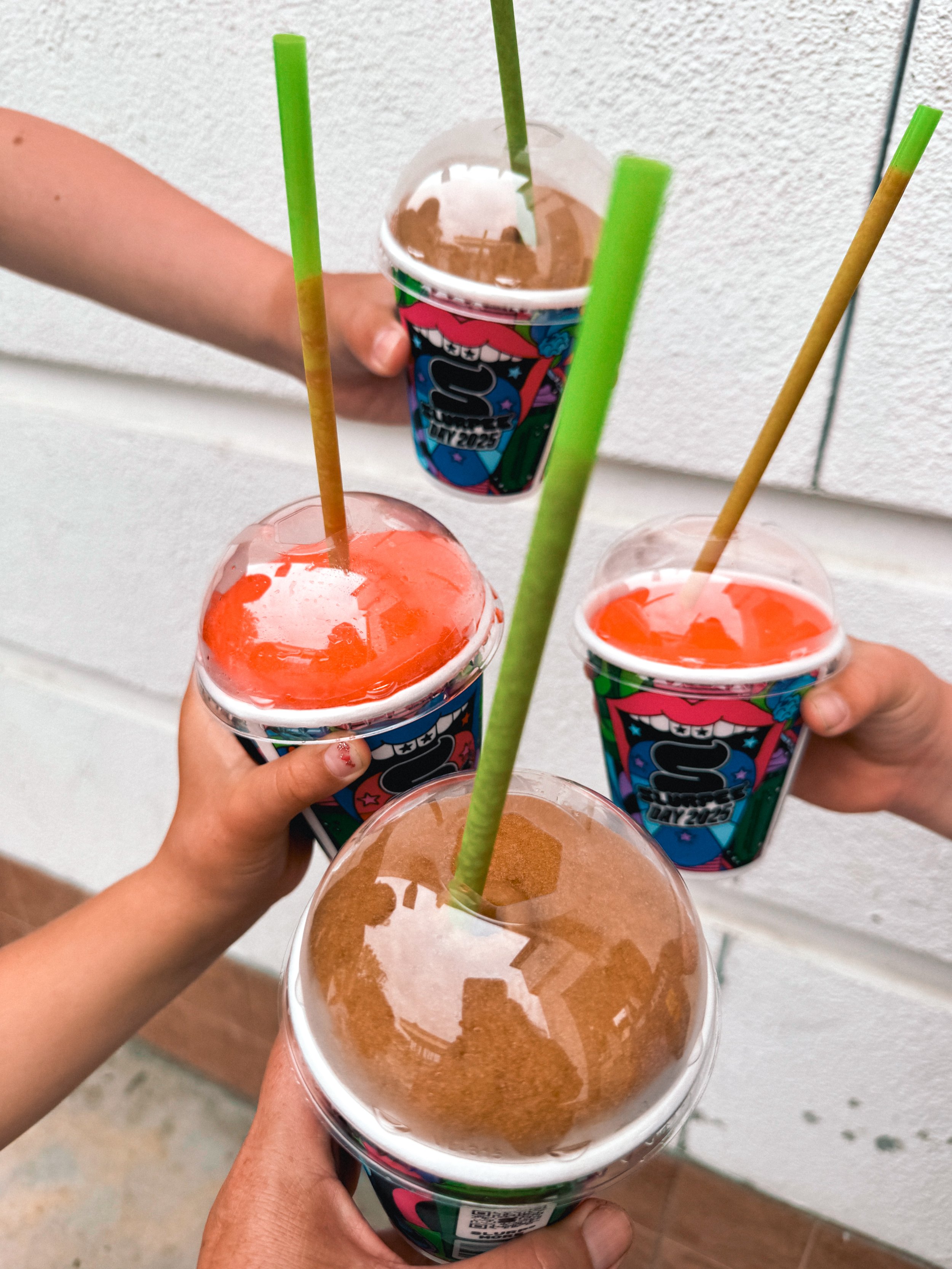Multiple people holding cups of frozen beverages, likely ice slushies, in colorful cups with straws, against a white wall.