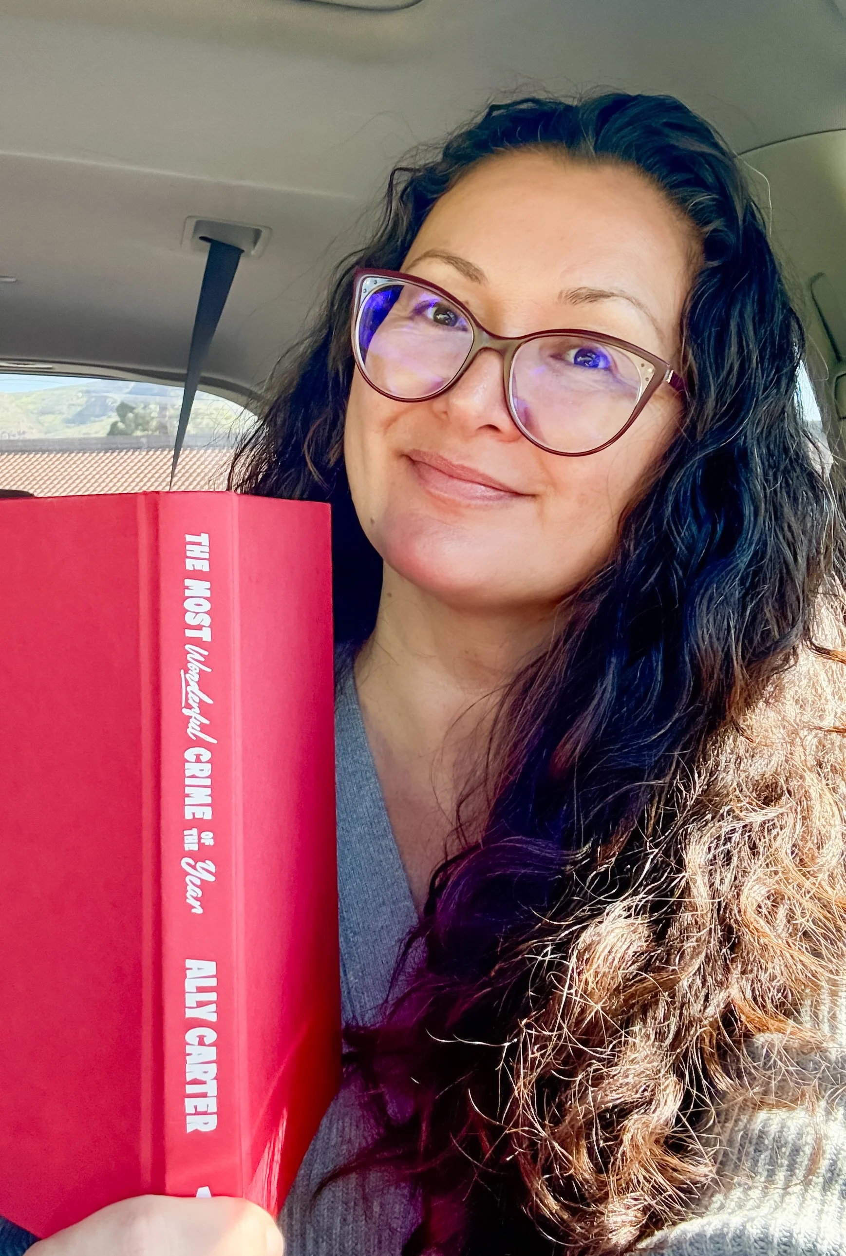 A woman with curly hair and glasses sitting inside a car, holding a red book titled 'The Most Important Crime of All'. The background shows a landscape with houses and trees through the car window.