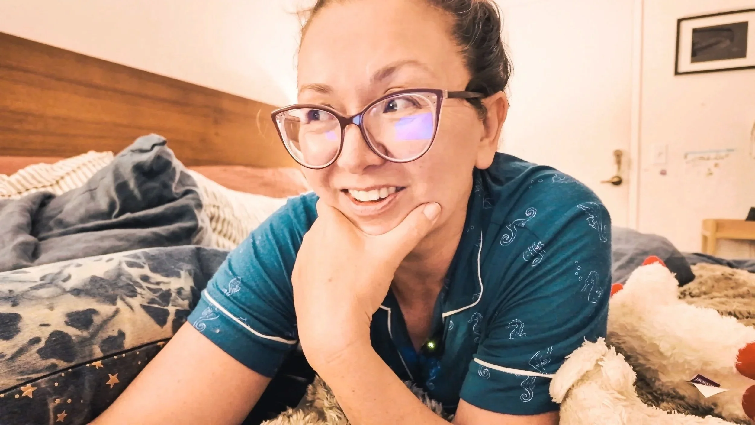 A woman with glasses and hair tied up lying on a bed, smiling, and resting her chin on her hand, with a dog next to her.
