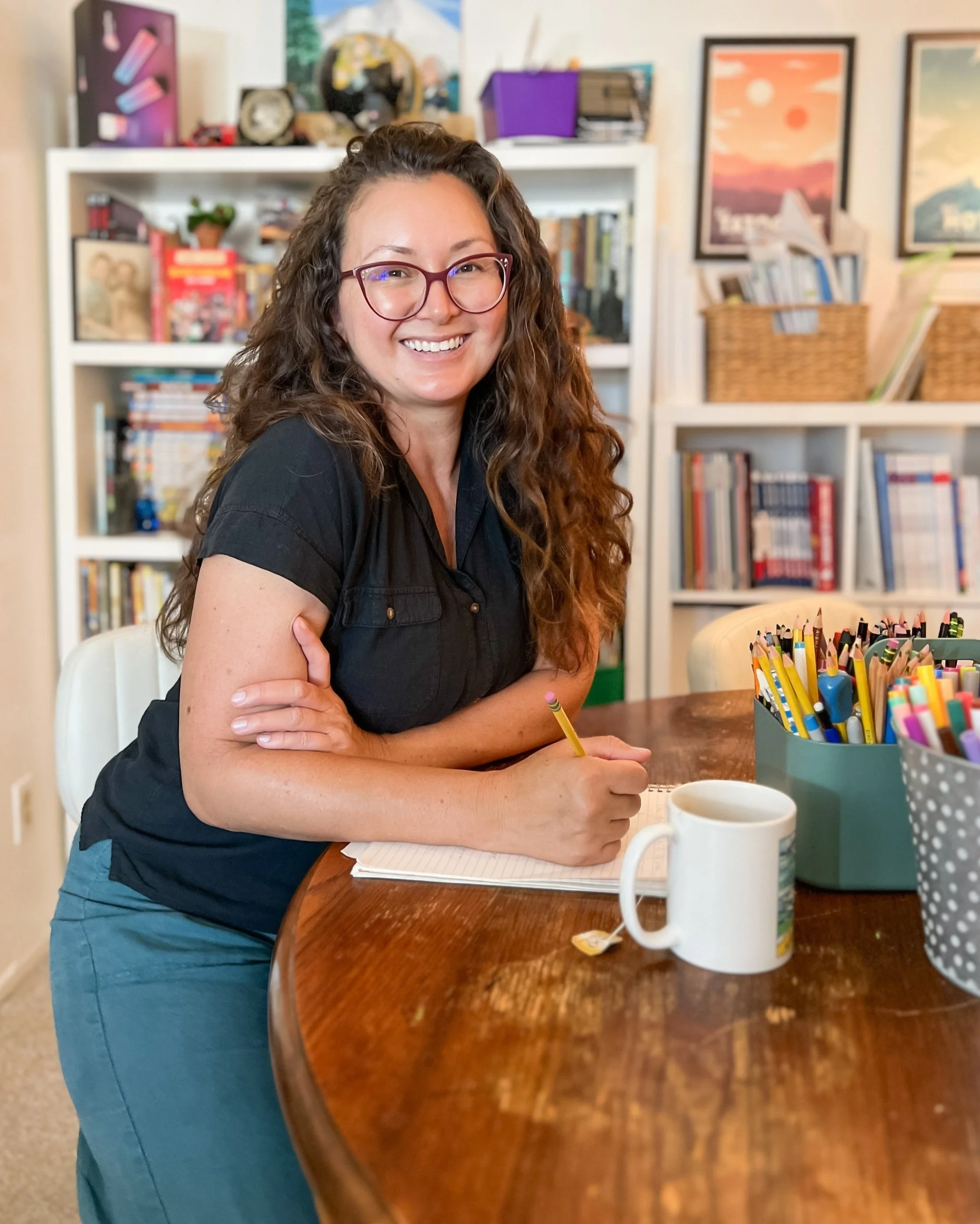 A woman with long curly hair, glasses, and a black shirt is sitting at a wooden table, smiling, with her arms crossed. The table has a cup, some notebooks, and a container of colored pencils. In the background, there are white bookshelves filled with books and decorations, with framed artwork on the wall.
