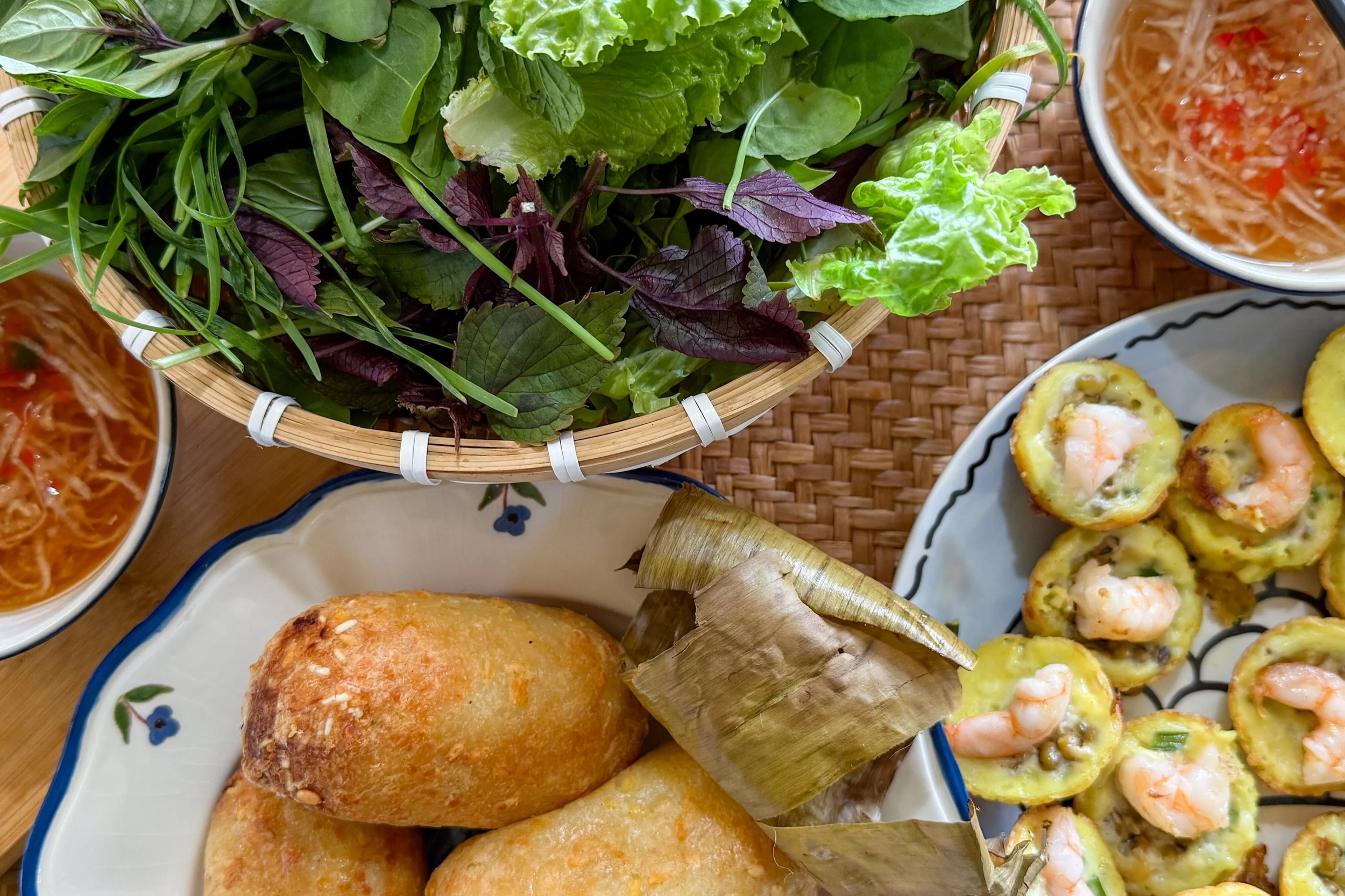 A basket of mixed leafy greens including purple and green lettuce, basil, and other herbs, surrounded by bowls of shrimp and vegetable dishes, and a plate of fried food on a woven placemat.