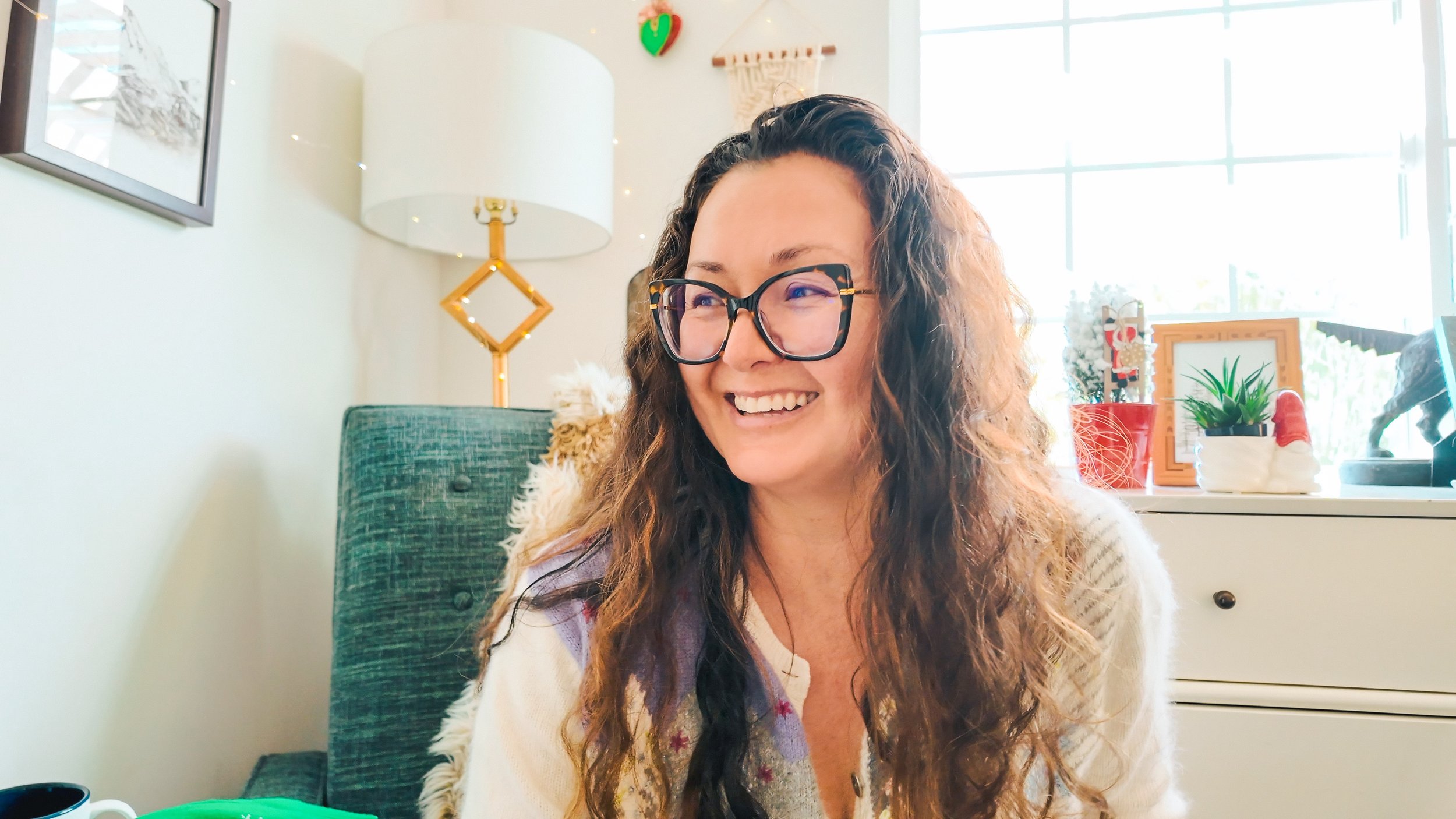A woman smiling and wearing glasses sitting in a well-lit room with holiday decorations, a teal armchair, and a white cabinet with plants and festive items.