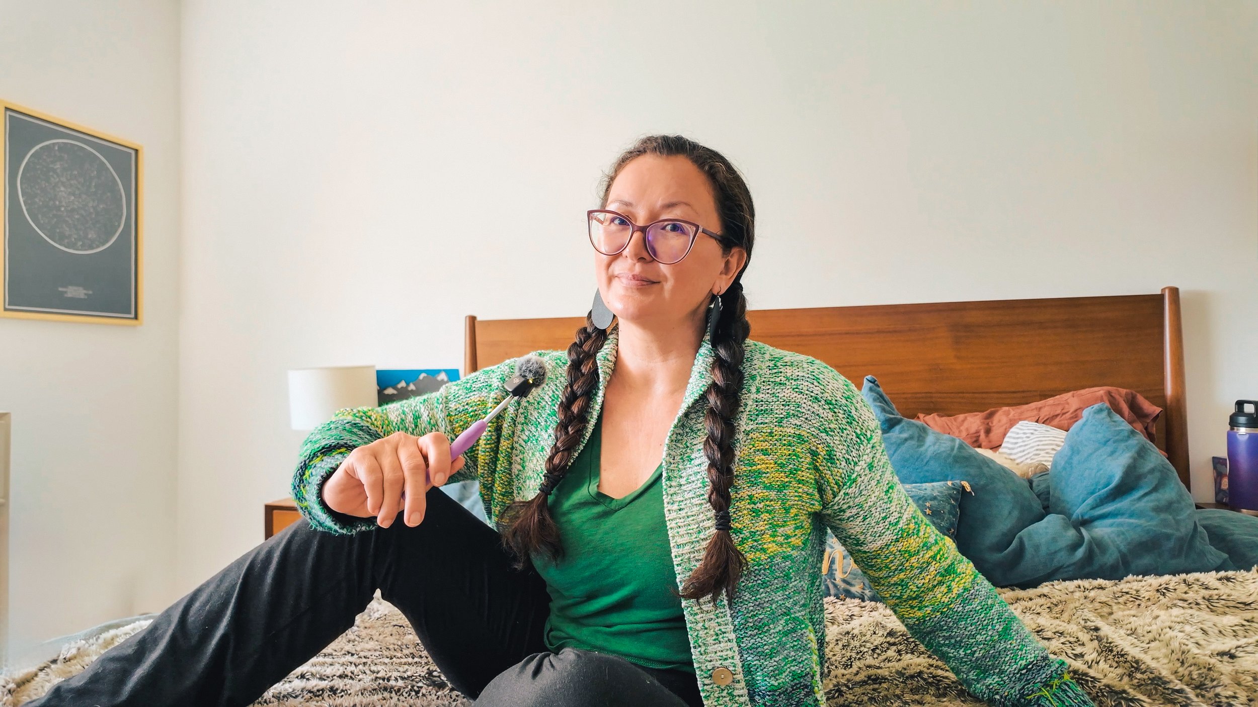 Woman with long braided hair and glasses sitting on bed, holding a mini microphone, smiling.