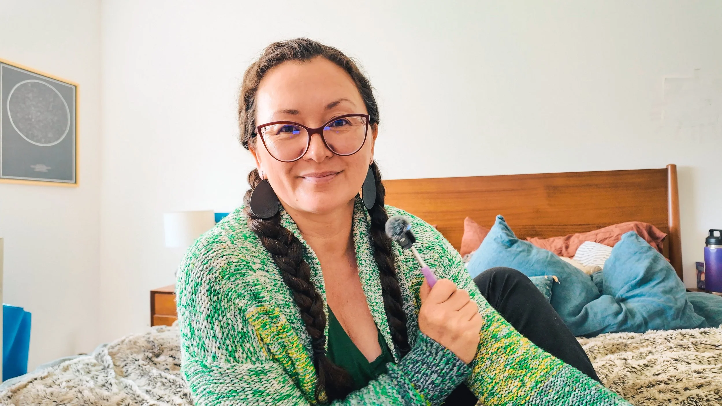 A woman with glasses, braided hair, and earrings sitting on a bed, holding a microphone, in a bedroom with blue pillows, brown headboard, and framed art on wall.