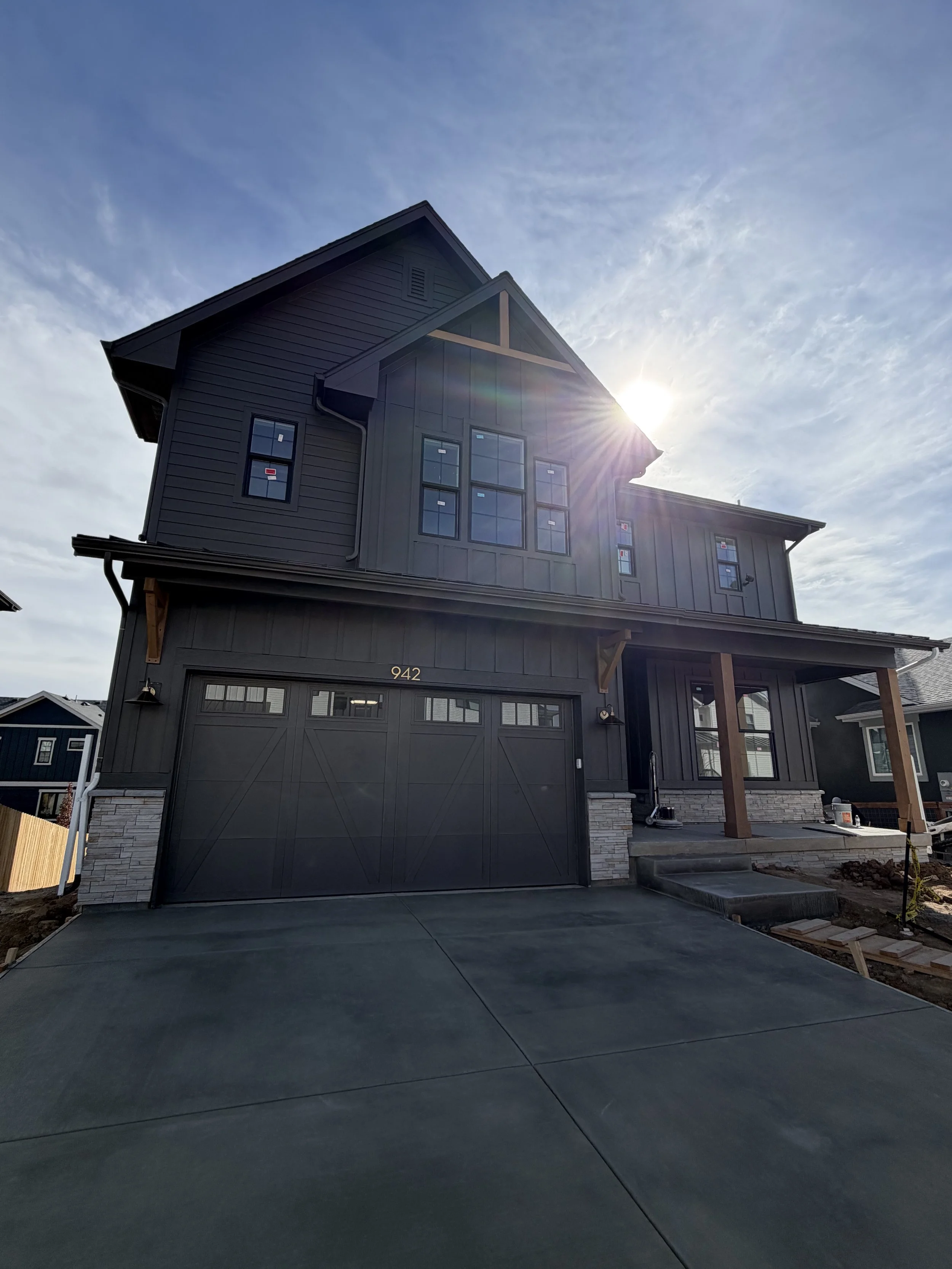 New two-story house with dark gray siding, stone accents, and a large garage door. The house has multiple windows and is under construction with some materials and tools visible outside.
