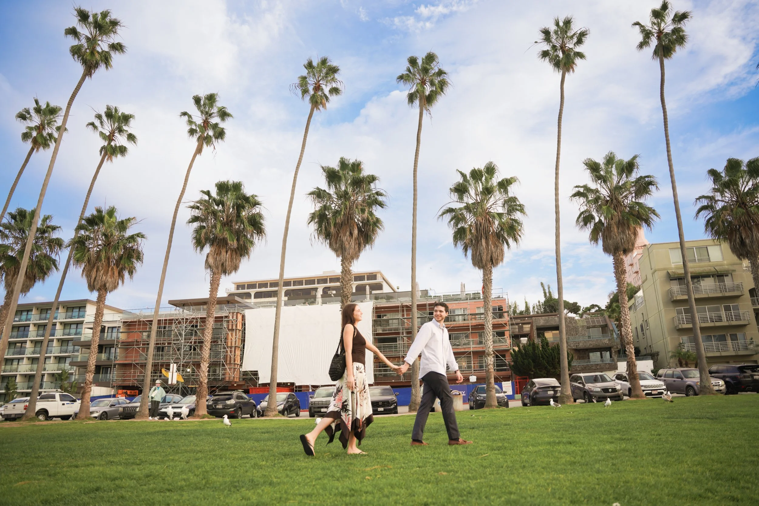 Noah and Charlotte walking hand in hand through Scripps Park at La Jolla Cove after their surprise proposal, sunset light over the Pacific behind them.