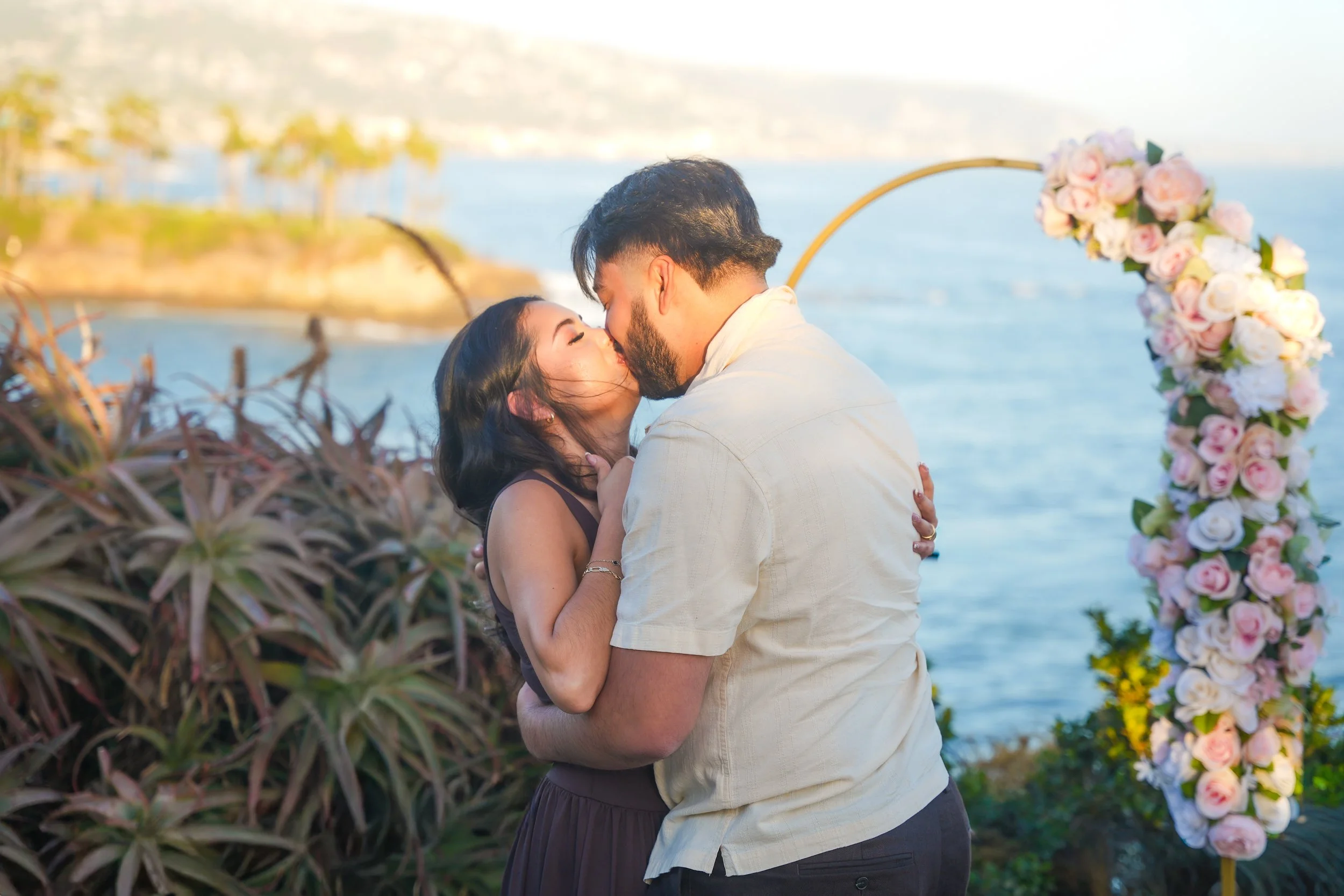 Couple sharing their first kiss after a surprise proposal in Laguna Beach beside a floral arch overlooking the Pacific Ocean, captured by a Southern California proposal photographer.