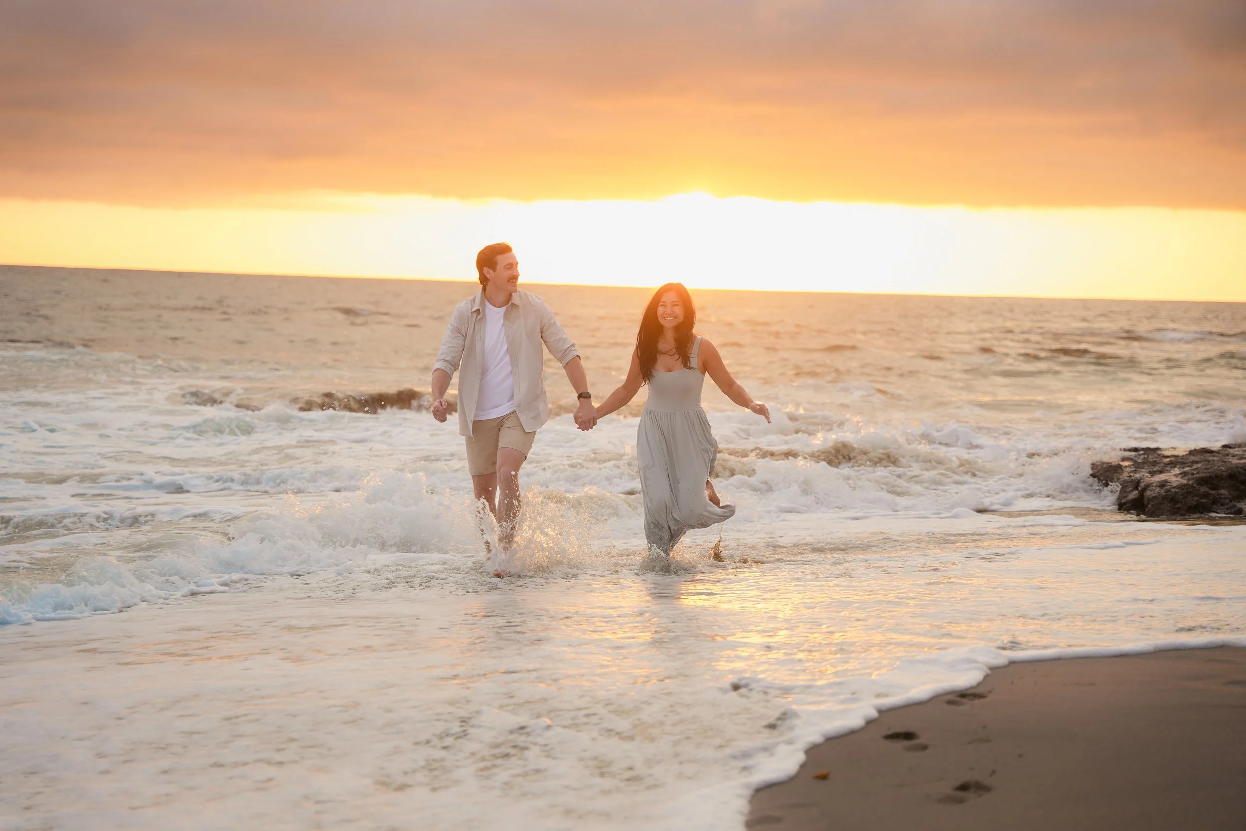 Nick and Tam running through the ocean water at sunset during their post-proposal engagement photos at Victoria Beach in Laguna Beach, California.