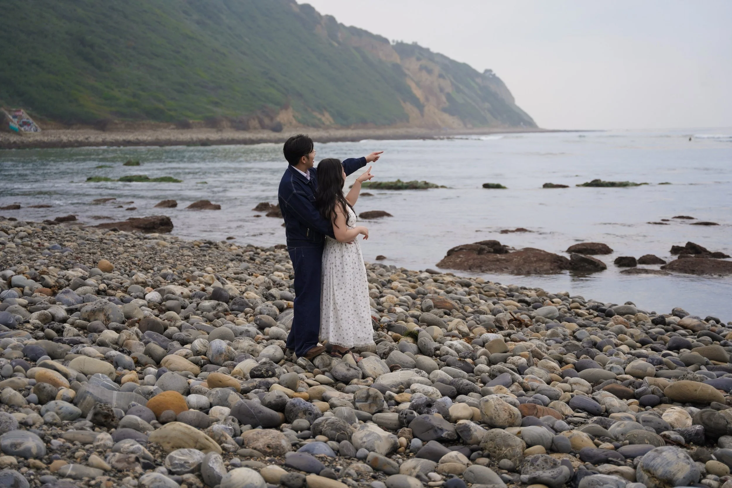 A candid pre-proposal moment during a surprise proposal at Palos Verdes Estates, as Johnny and Phong explore the Bluff Trail shoreline with ocean cliffs and the Pacific Ocean behind them.