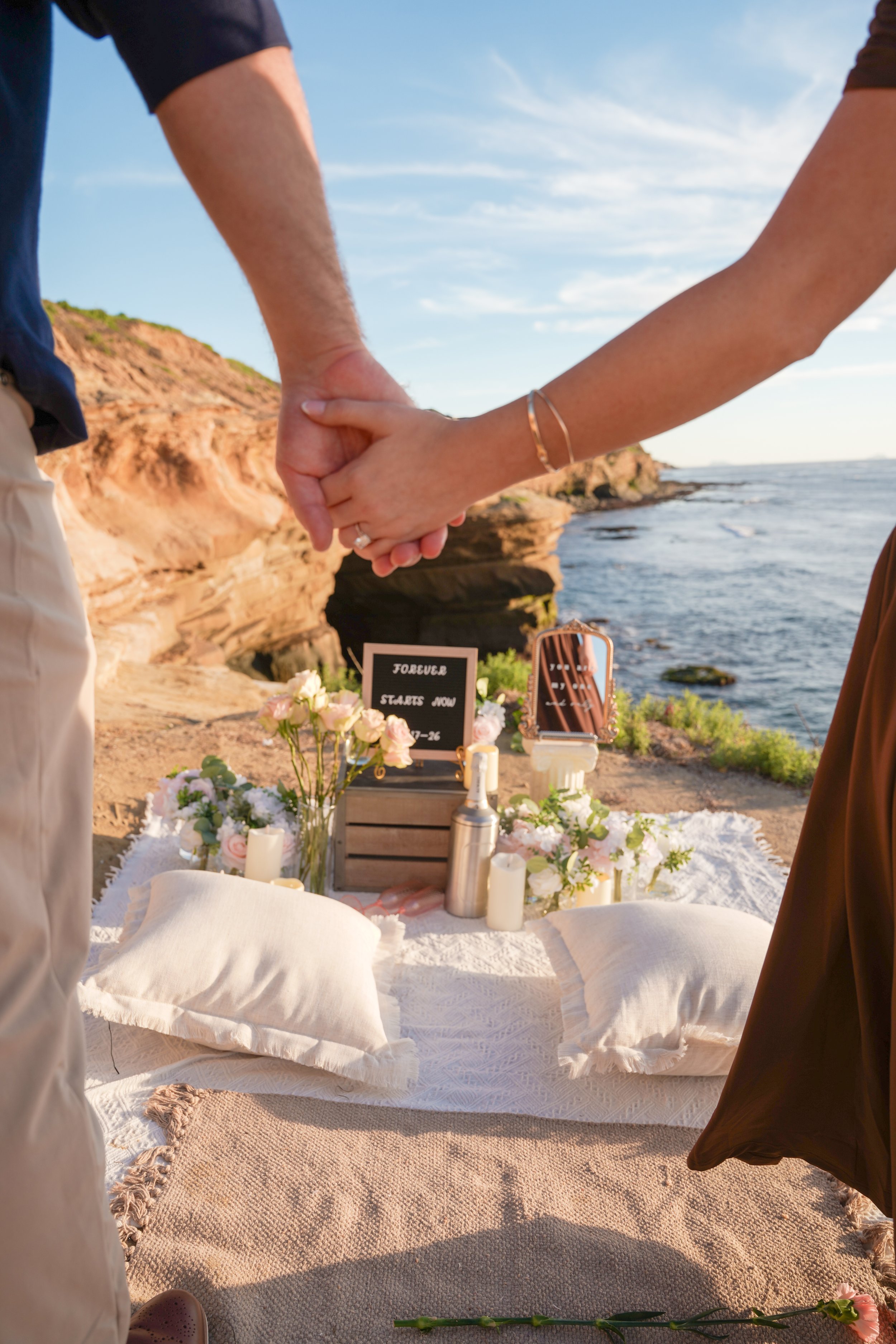 Minimal boho surprise proposal setup with neutral pillows, floral arrangements, and custom felt letter board overlooking a scenic outdoor location