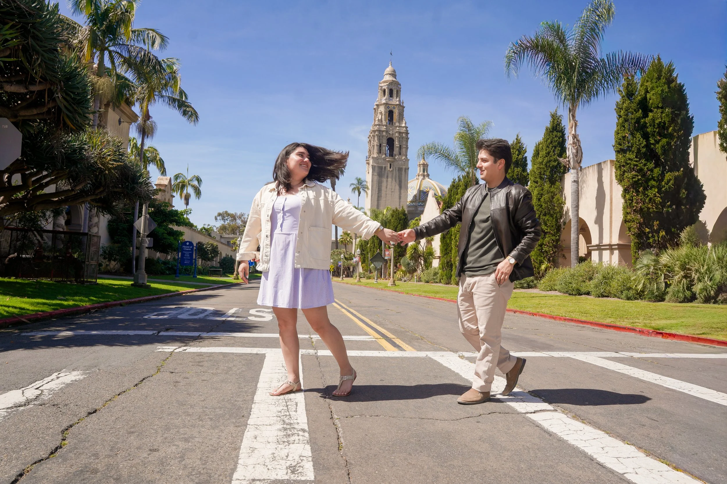 Couple walking across a Balboa Park walkway in San Diego after their proposal, with the iconic California Tower in the background, captured in natural light engagement photography