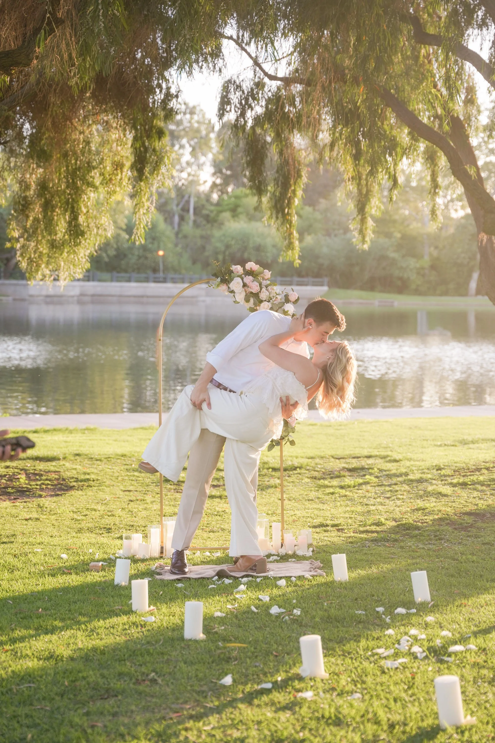 Luxury surprise proposal setup featuring a gold circular arch with soft pink and white floral accents, rose petal walkway, and candlelight at sunset, designed to elevate an intimate engagement moment