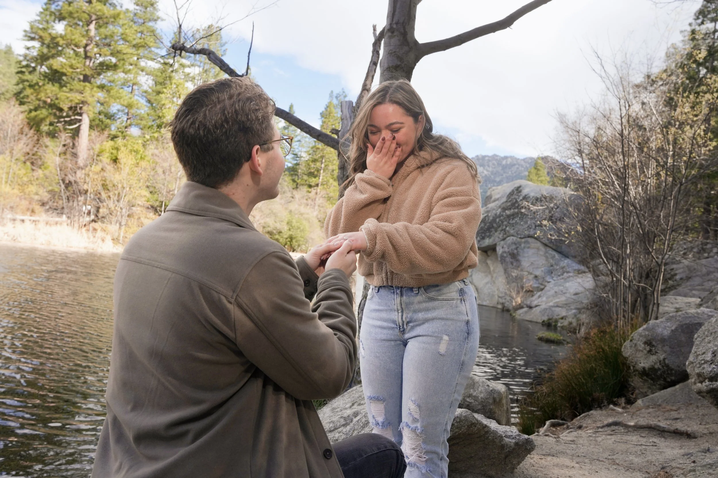 Man kneeling during a surprise proposal at Lake Fulmor in Idyllwild, asking his partner to marry him beside a peaceful forest lake surrounded by pine trees in the San Jacinto Mountains