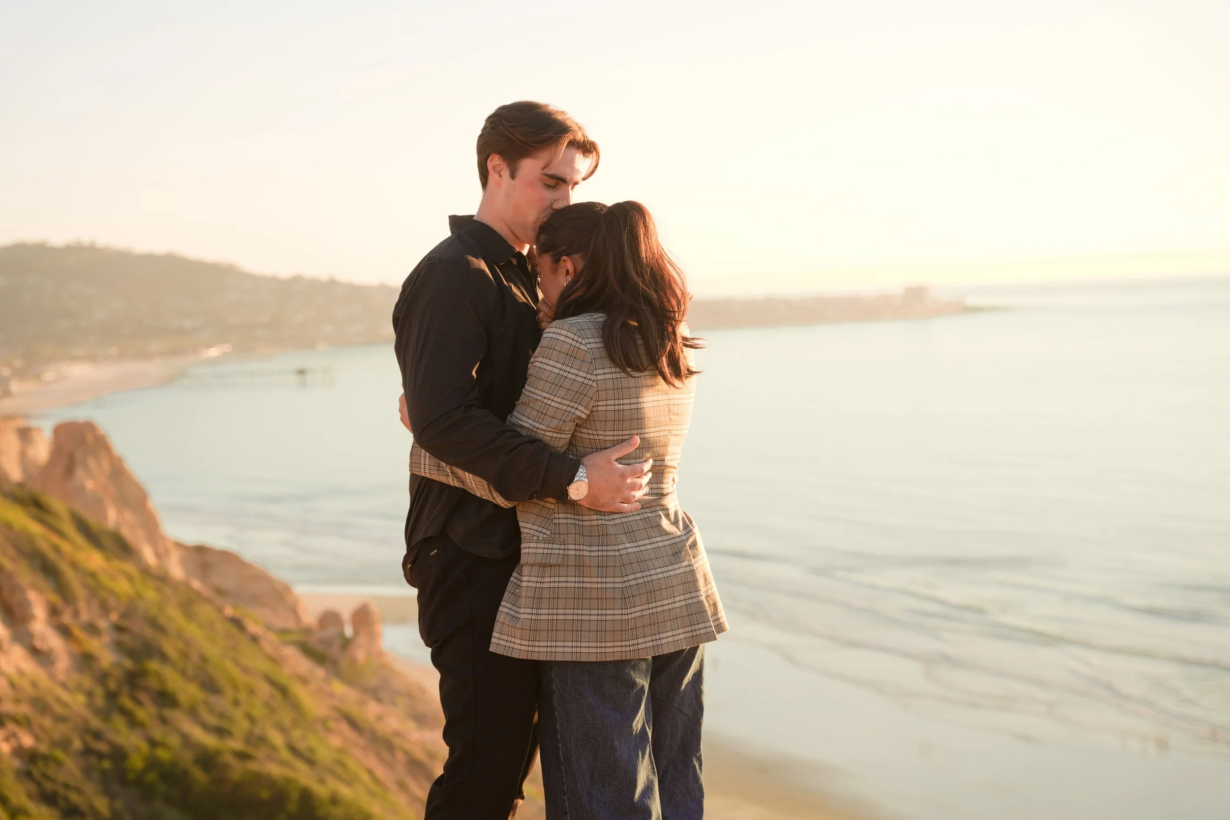 Couple embracing at Torrey Pines Gliderport during sunset, overlooking the Pacific Ocean cliffs, captured during a romantic engagement or post-proposal photoshoot in La Jolla.