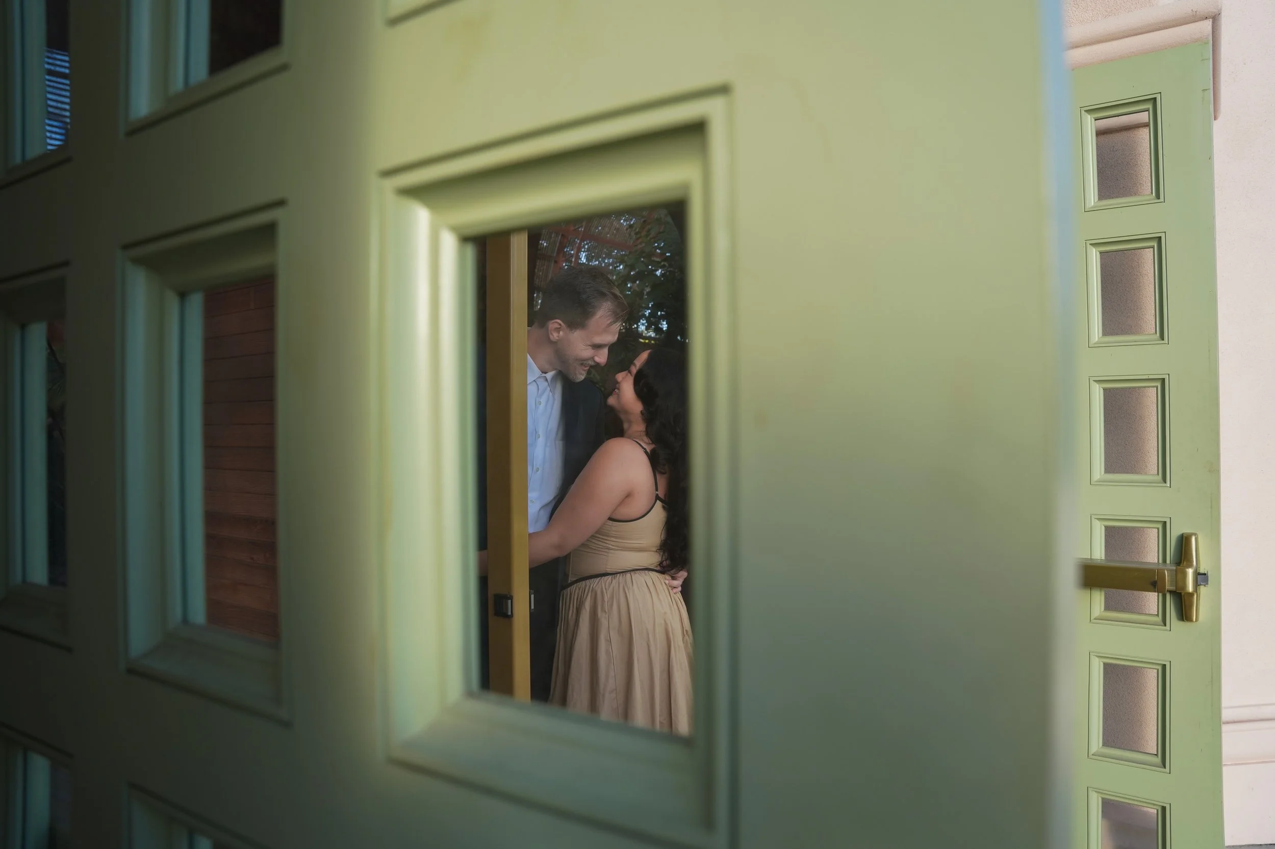 CJ and Dessa embracing inside the Botanical Building at Balboa Park during their San Diego surprise proposal, captured through a window for an intimate, candid moment.