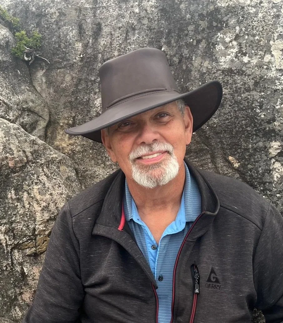 A man with a white beard and mustache wearing a wide-brimmed gray hat, a blue collared shirt, and a black jacket, smiling in front of a rocky background.