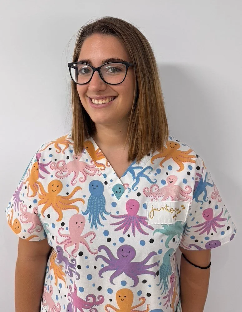 Mujer sonriendo con gafas, lleva uniforme con dibujos de pulpos en diferentes colores y un parche con la palabra 'juntos'.