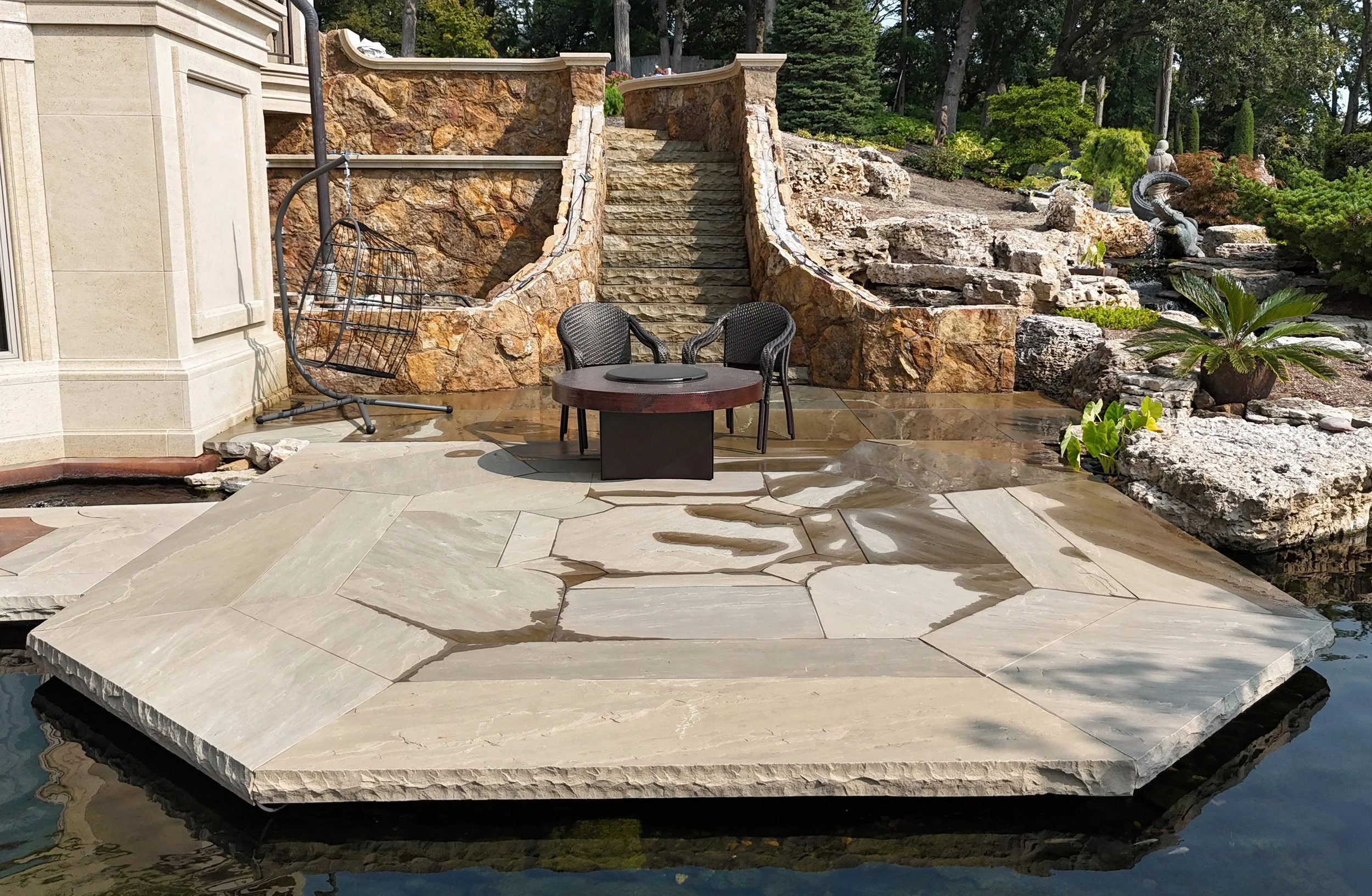 A backyard patio with a hot tub, stone stairs, two black chairs, a round table, a hanging chair, a rock waterfall feature, and lush greenery.