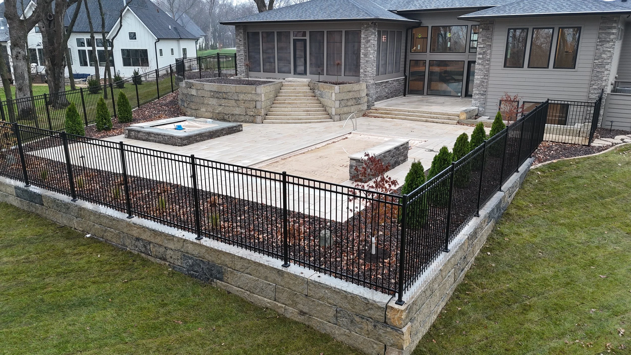 Backyard patio area with beige stone pavers, black metal fence, and landscaped garden beds with small evergreen shrubs.