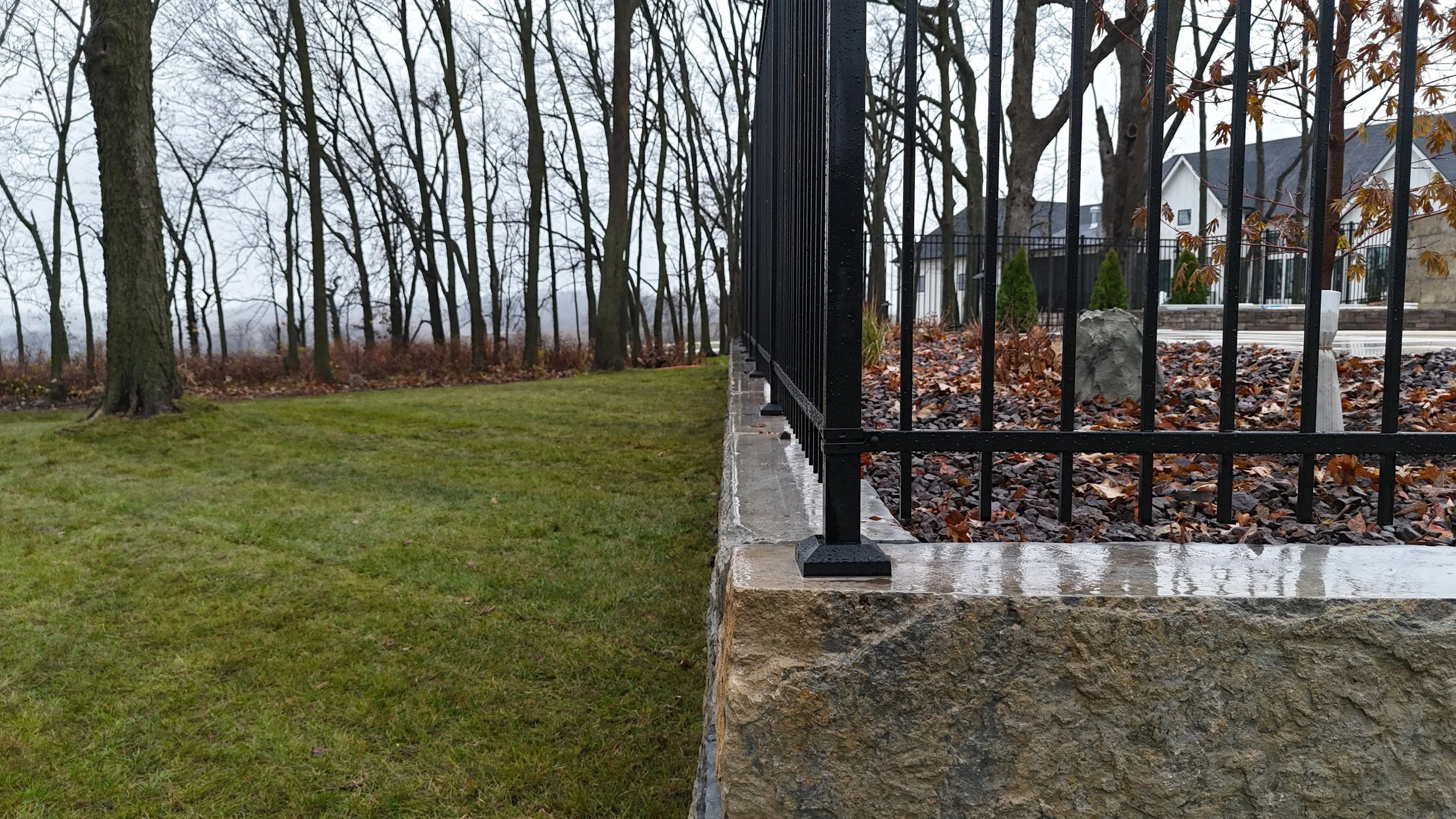 A close-up view of a black metal fence installed on a stone foundation, with a residential neighborhood in the background, trees, fallen leaves, and a grassy yard, on a cloudy day.