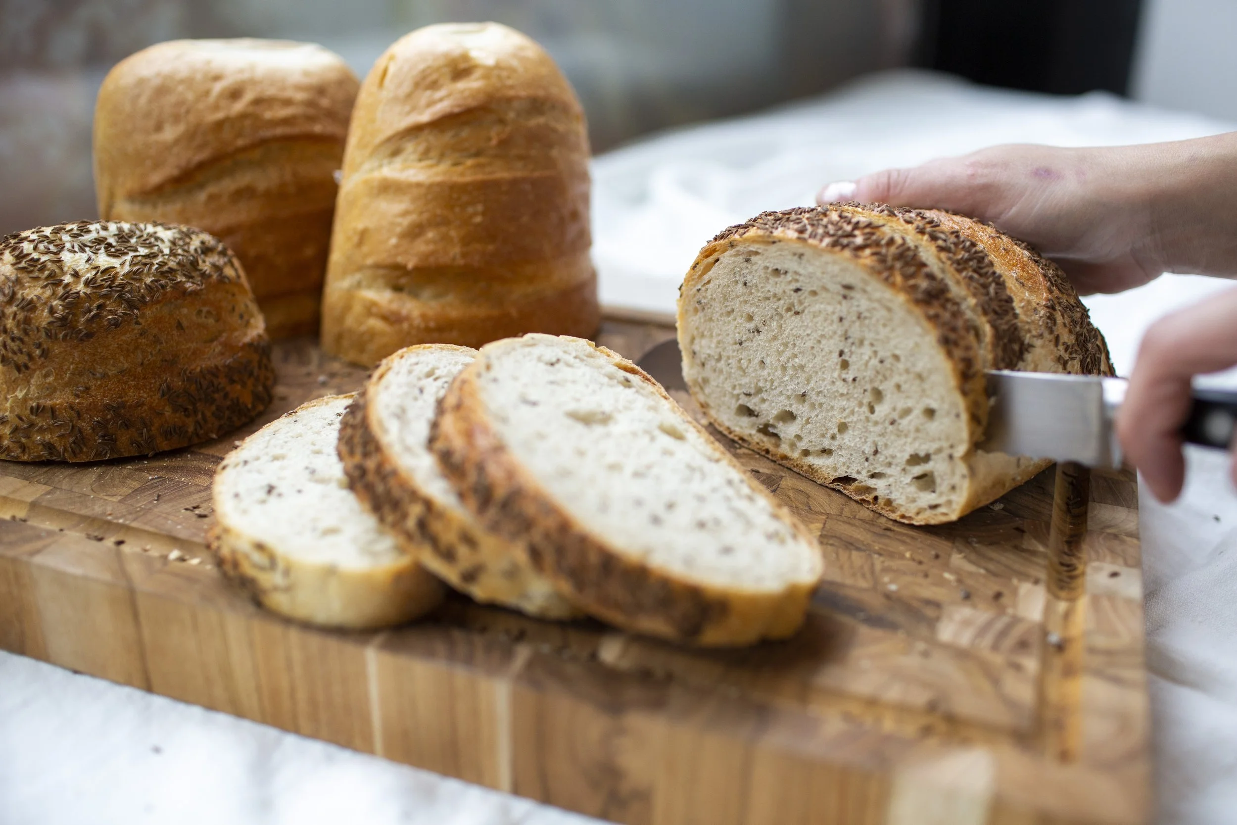 Wooden cutting board with slices of cut bread in the foreground, and a seeded rye being sliced in the background. 