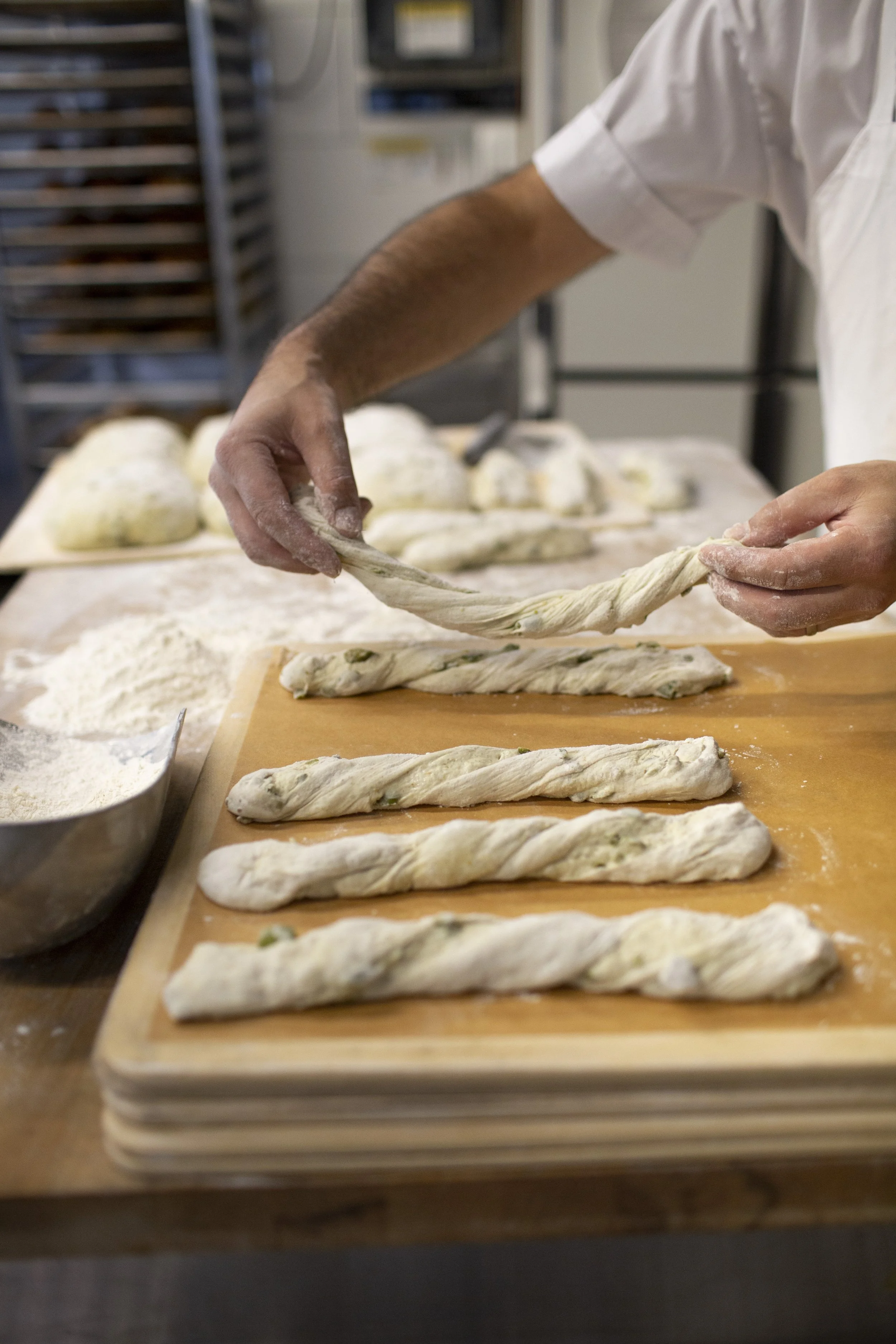A chef in a white jacket is twisting and laying out pre-baked, rolled olive sticks in the foreground; in the background are flour vessels, dough waiting to be baked and a baking rack. 
