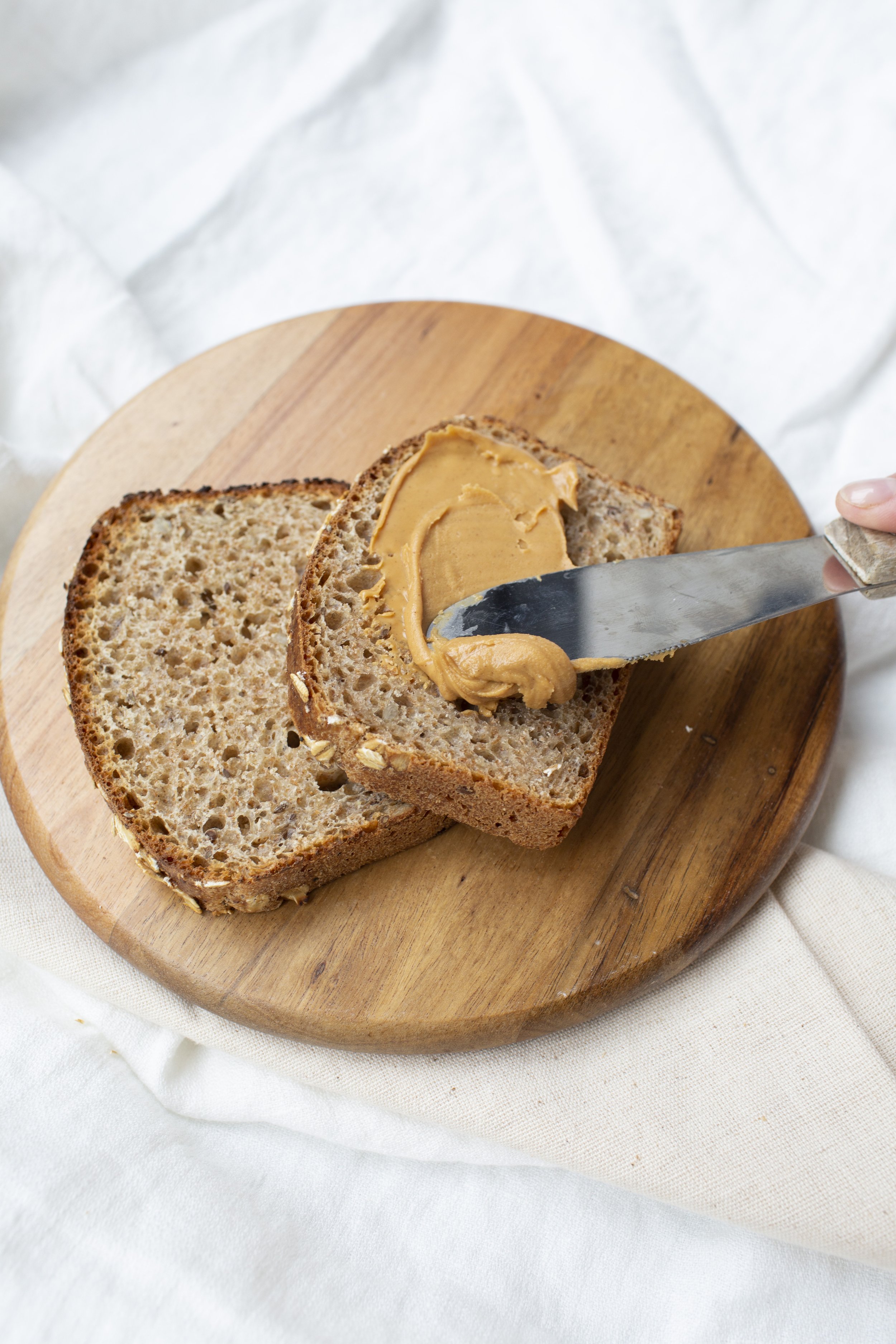 Sliced spelt bread on a round cutting board, with spreading peanut butter lathered on the top piece.