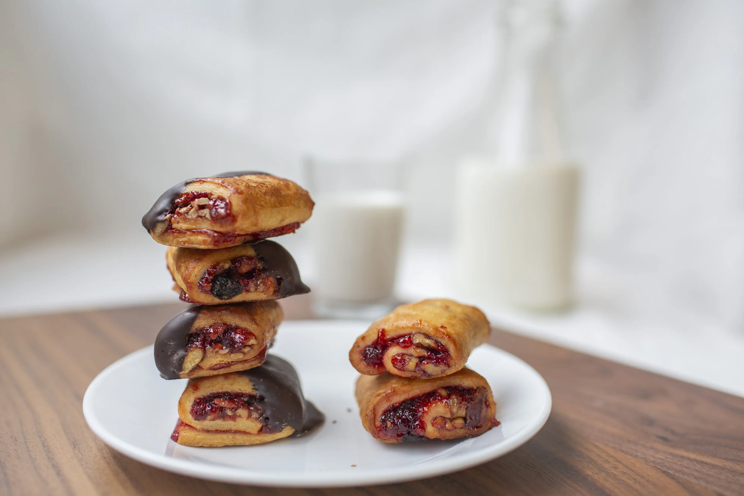Two stacks of rugelach cookies dipped in chocolate on a white plate; milk in the background.