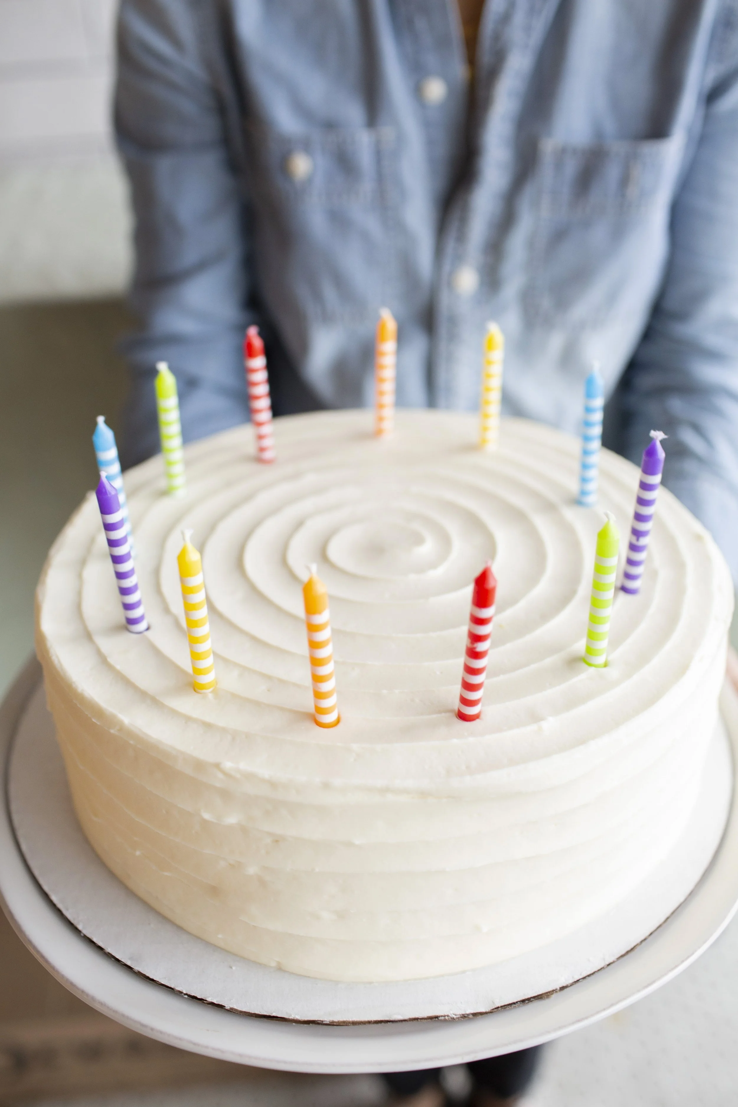 White buttercream frosted cake, adorned with rainbow colored candles, held by a baker in a denim blue jean shirt.