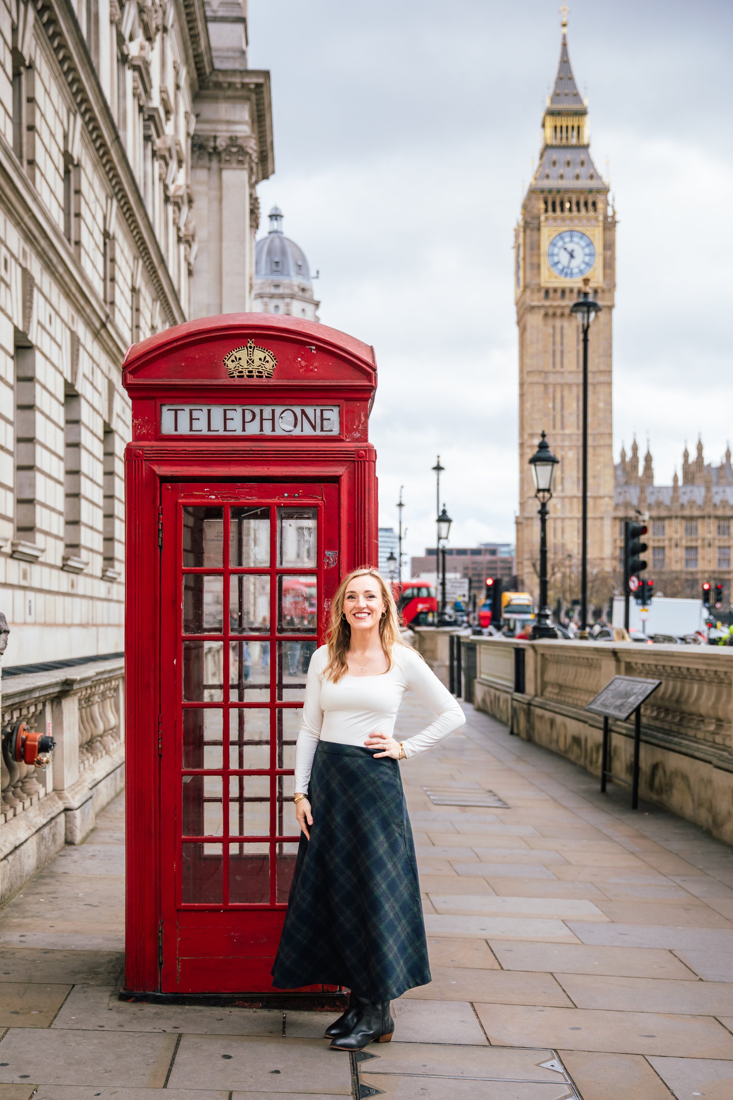 A woman standing next to a red British telephone booth on a city street in London, with Big Ben clock tower in the background.