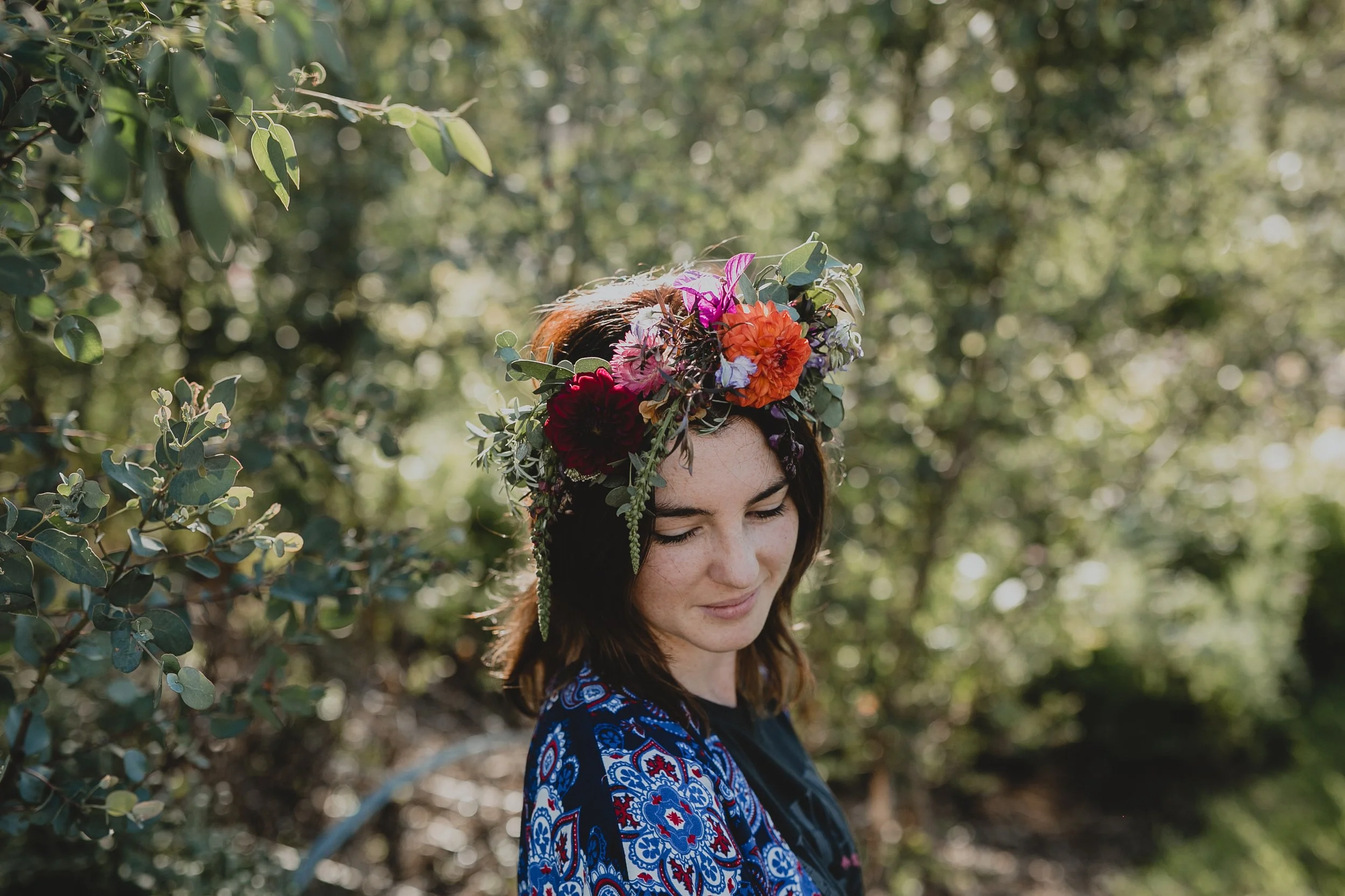 Emma wearing a floral crown made of colorful flowers including a big golden Marigold, standing outdoors in a wooded area.