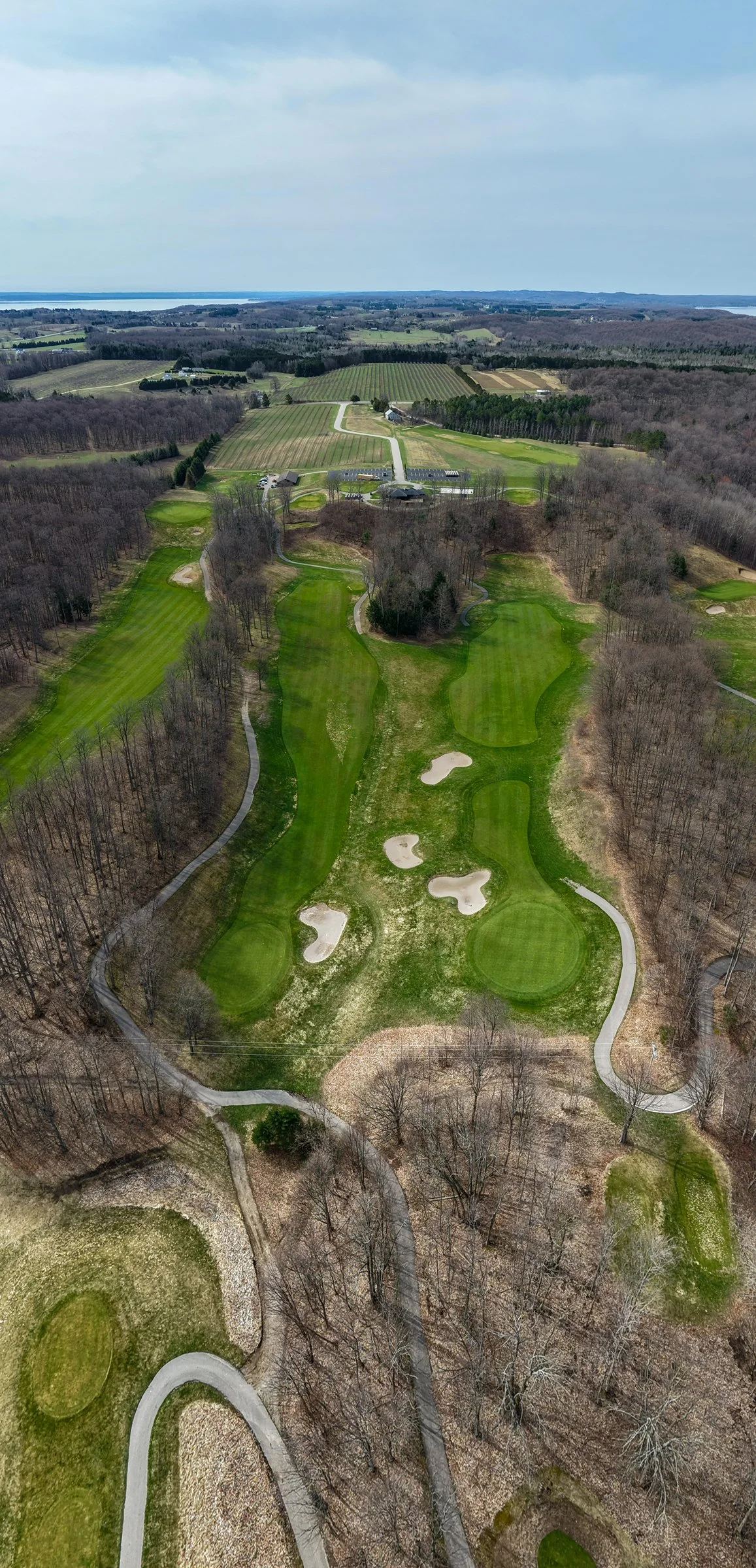 Aerial view of a golf course with green fairways, sand bunkers, and surrounded by leafless trees, with farmland and distant water in the background.
