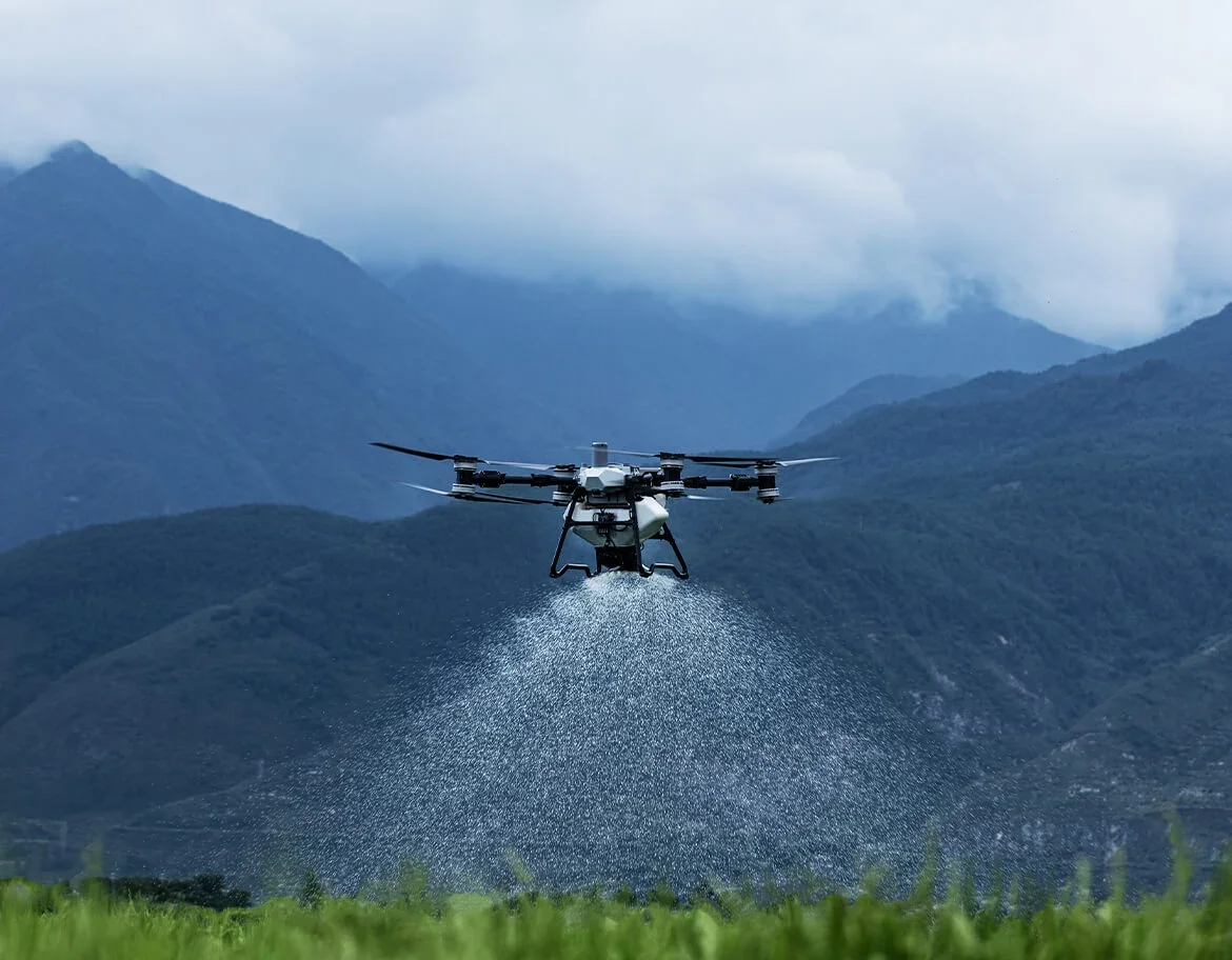A drone spraying water over a green field with mountains in the background.