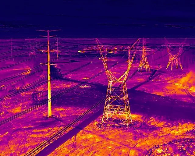 Infrared image of electrical transmission towers and power lines over a landscape with fields and sky.
