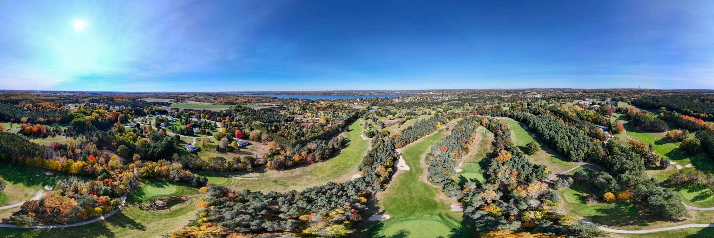 Aerial view of a golf course surrounded by trees with fall foliage, residential houses, and a body of water in the background under a clear blue sky.