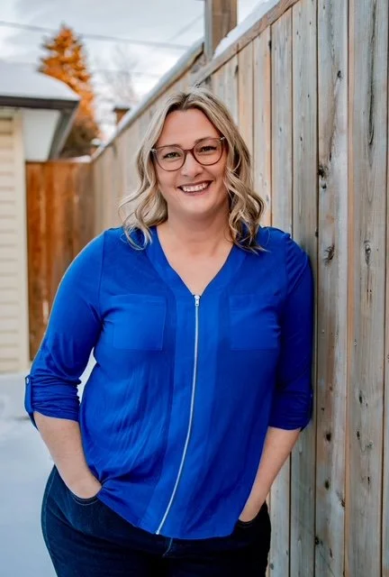 A woman with blonde hair and glasses smiling, wearing a blue blouse, standing outdoors next to a wooden fence.