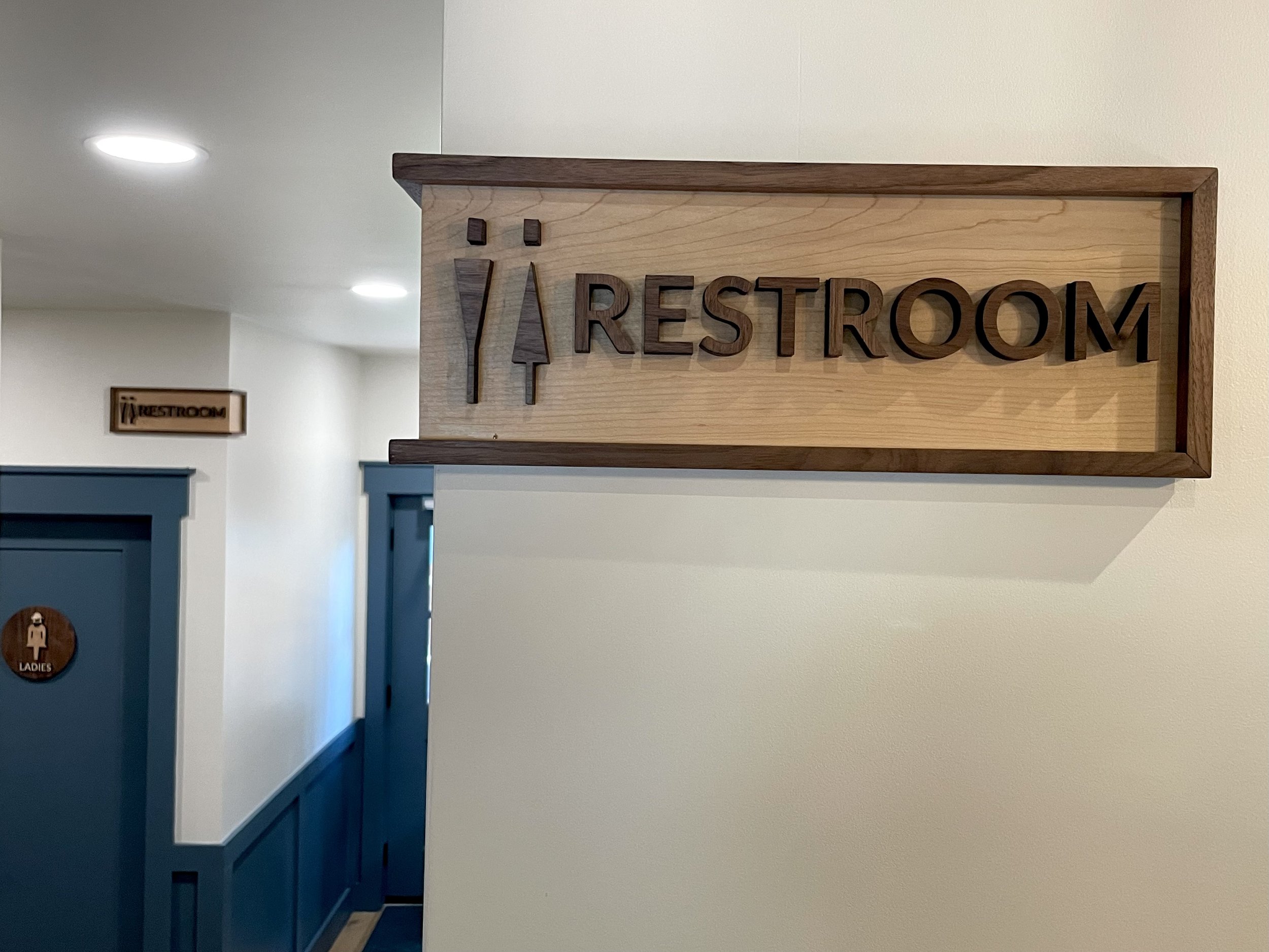 A Clear Path: Both wall signs in place, one at the hallway entrance and the second at the restroom corner. The Ladies door sign is also visible, creating a cohesive visual guide throughout the corridor.