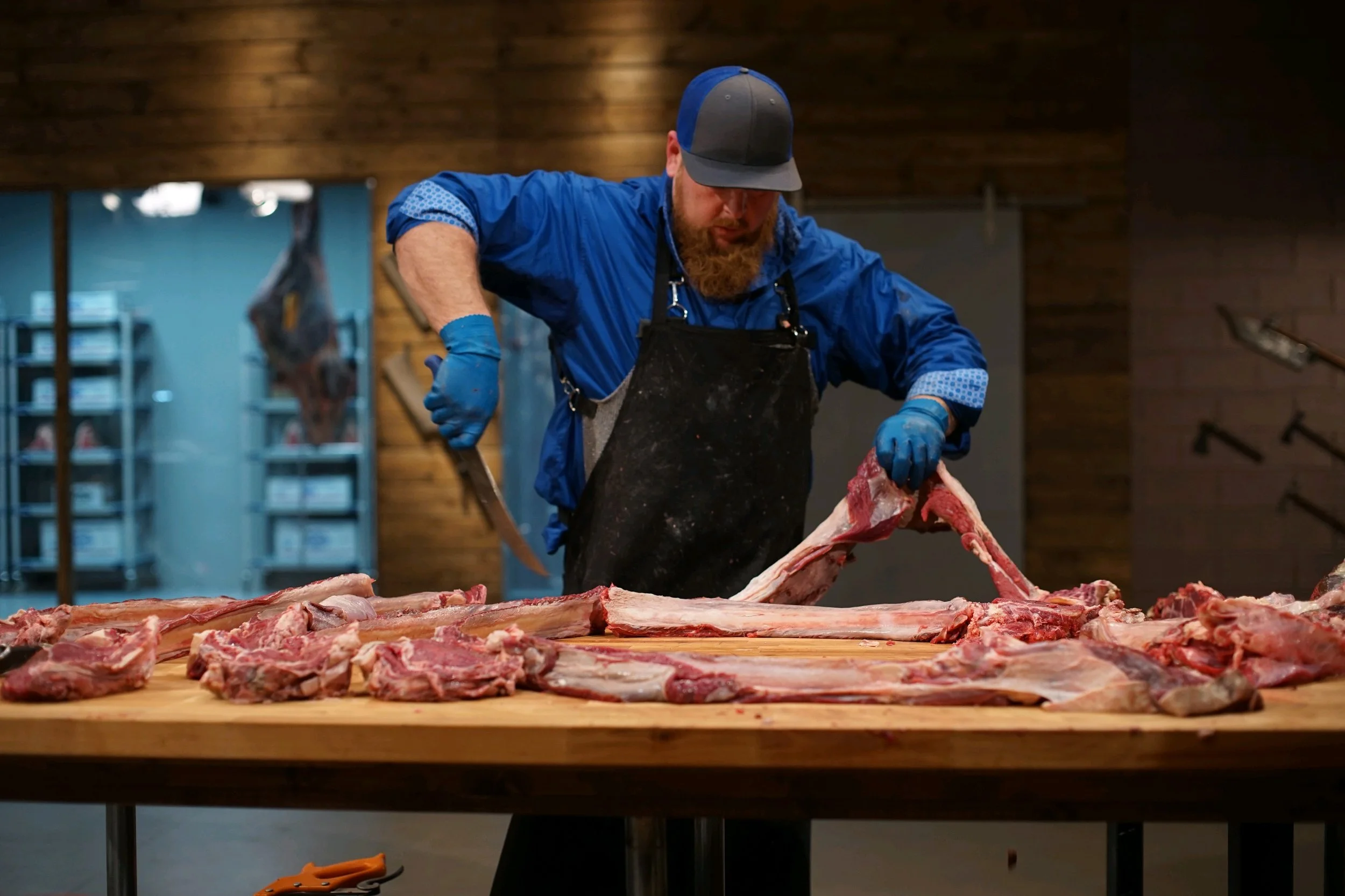 A man with a beard wearing a blue jacket, gray cap, and blue gloves butchering a large piece of meat on a wooden table, with additional meat pieces spread across the surface, in a room with wooden walls and shelves in the background.