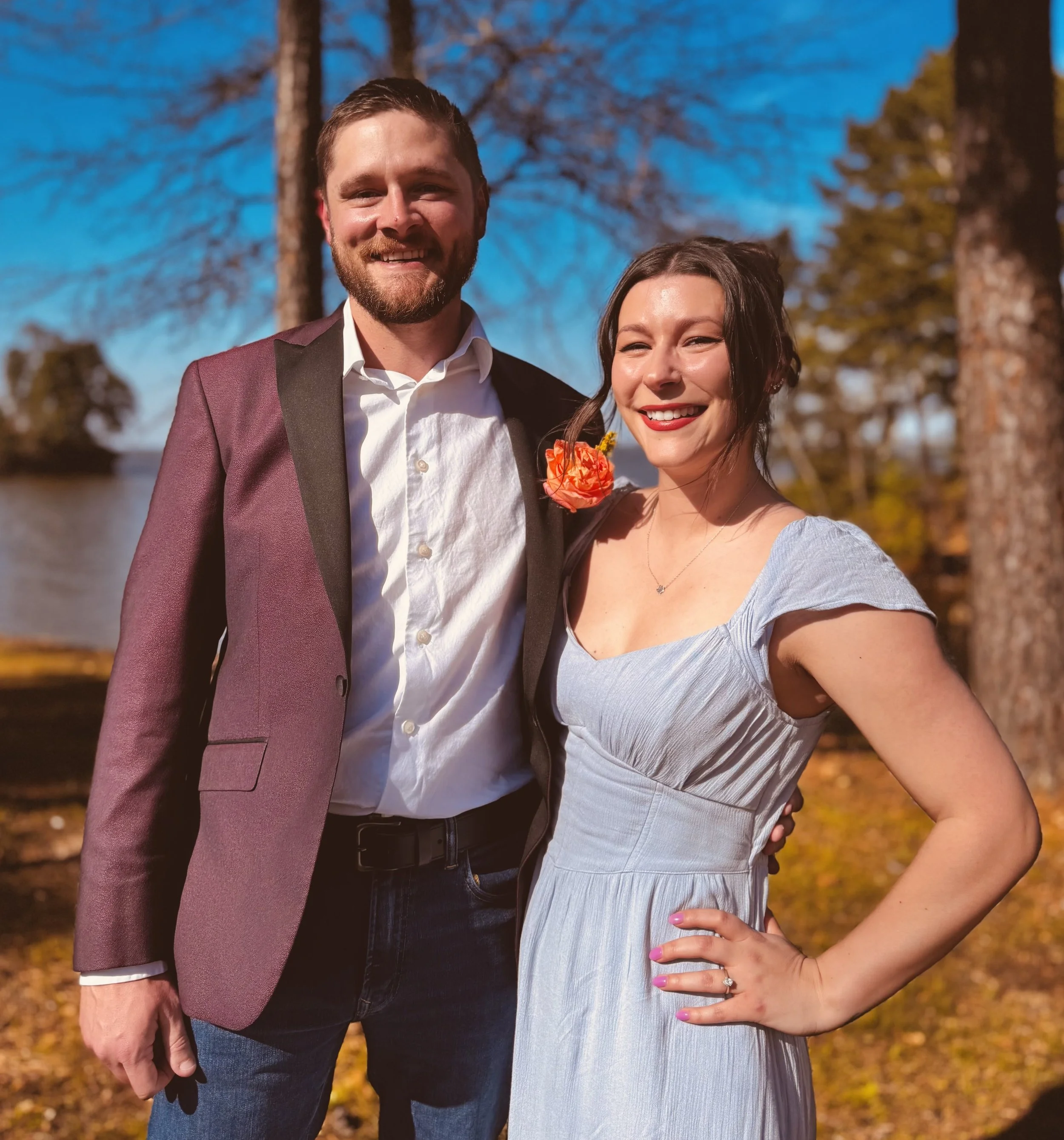 A smiling couple standing outdoors in front of trees and a body of water, dressed for a special occasion, with the man in a suit and the woman in a light gray dress.