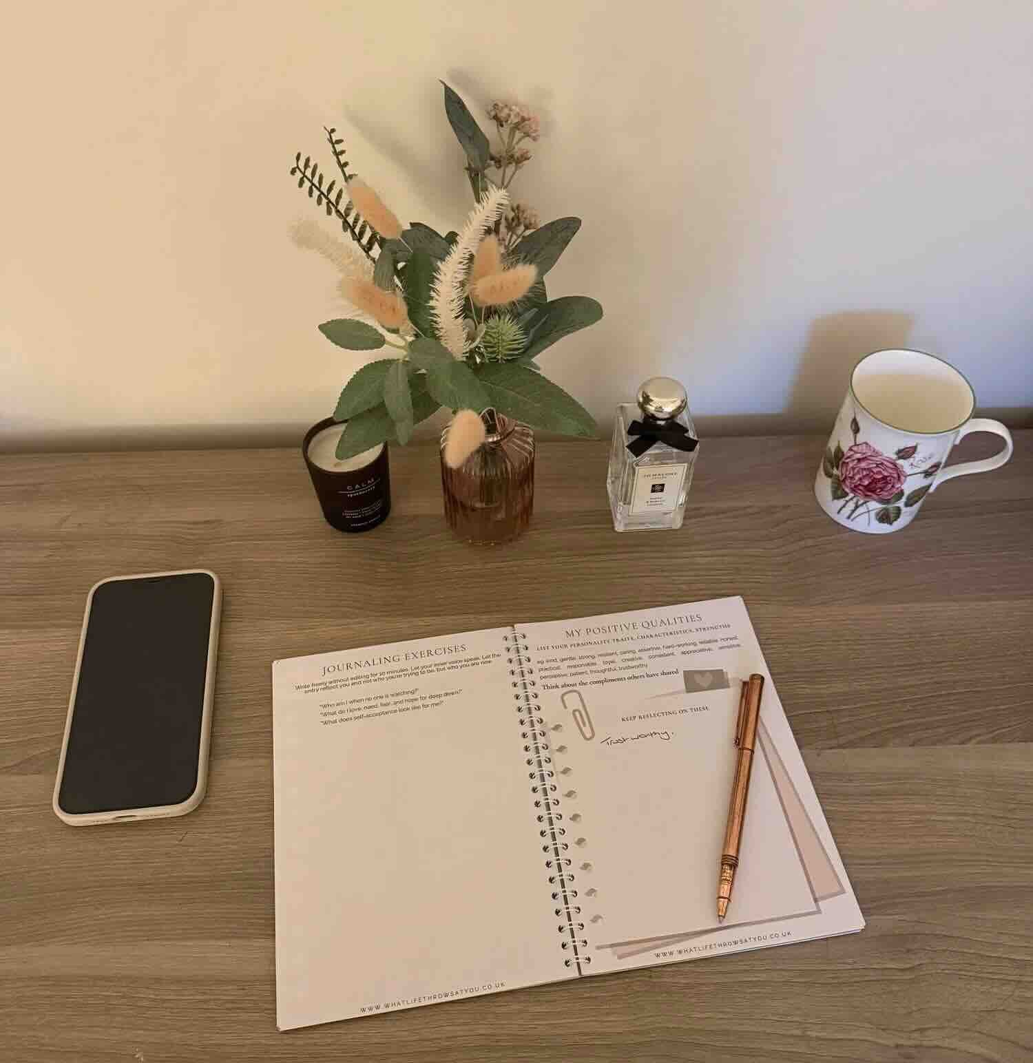 Open workbook on a dest with a plant perfume iphone coffee cup and candle and pen