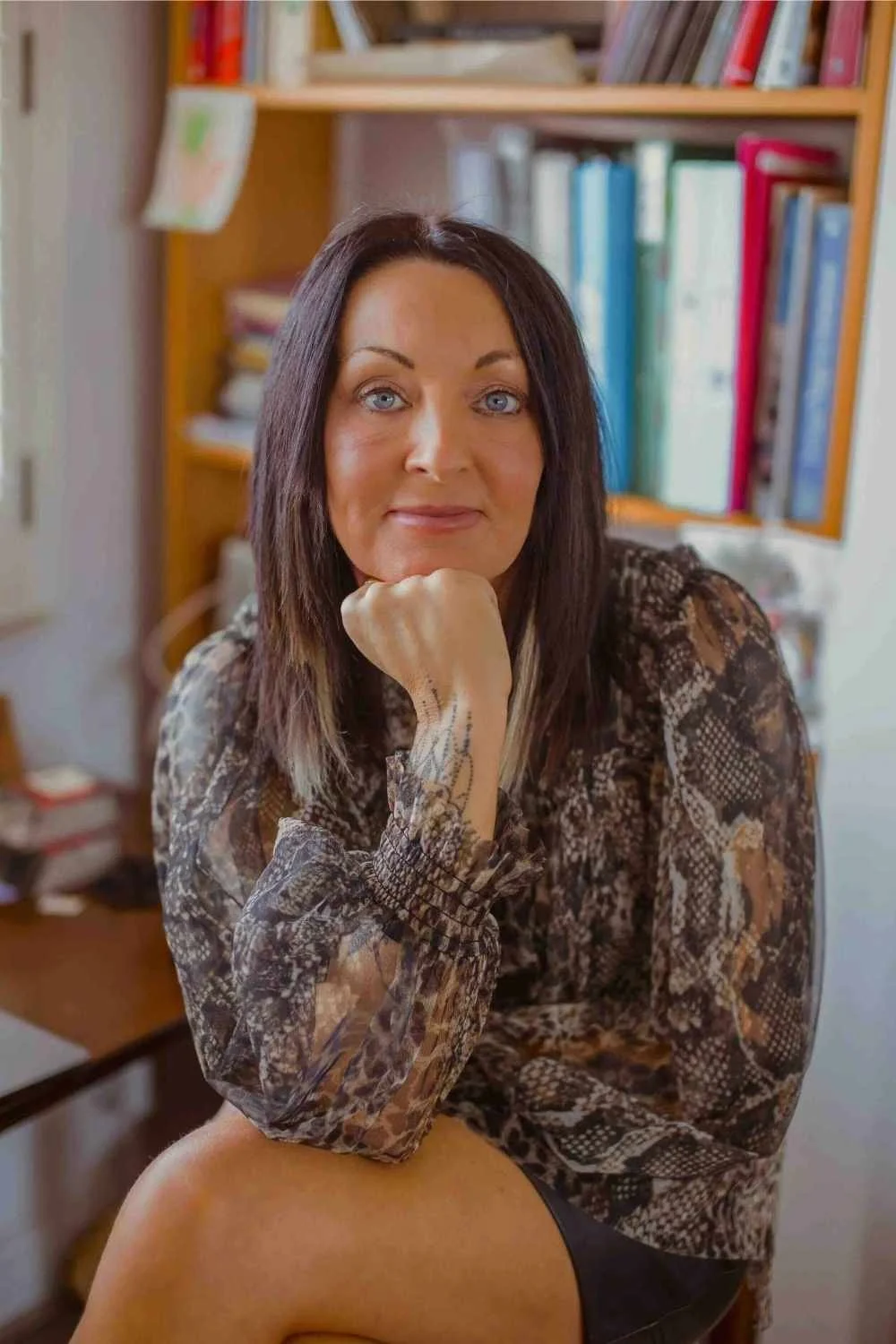 Photo of the author Andrea Bevan sat in front of a book shelf crossed legged leaning on her wrist